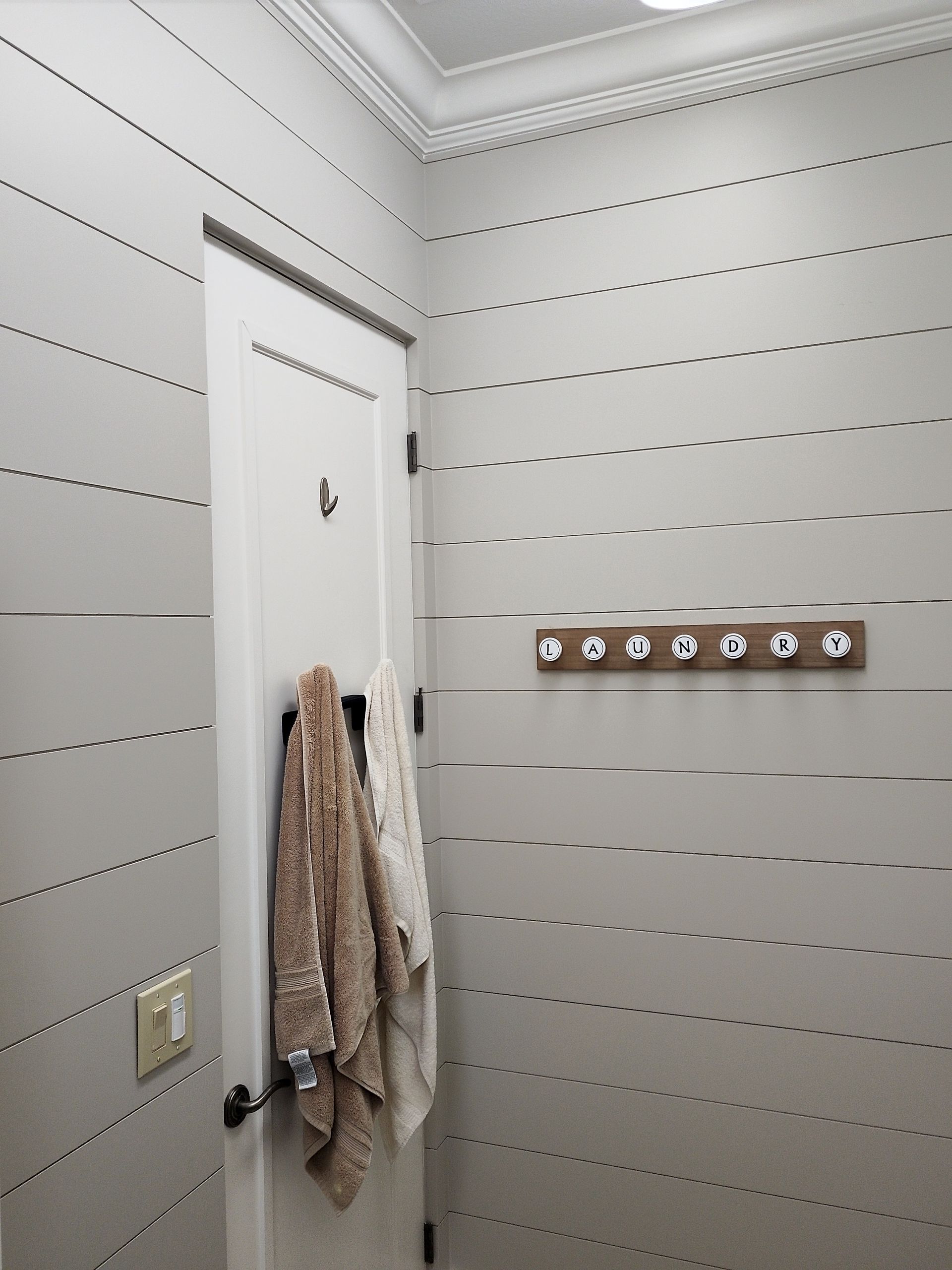 Bathroom corner with light grey horizontal panel walls, white door, towels, and coat rack.
