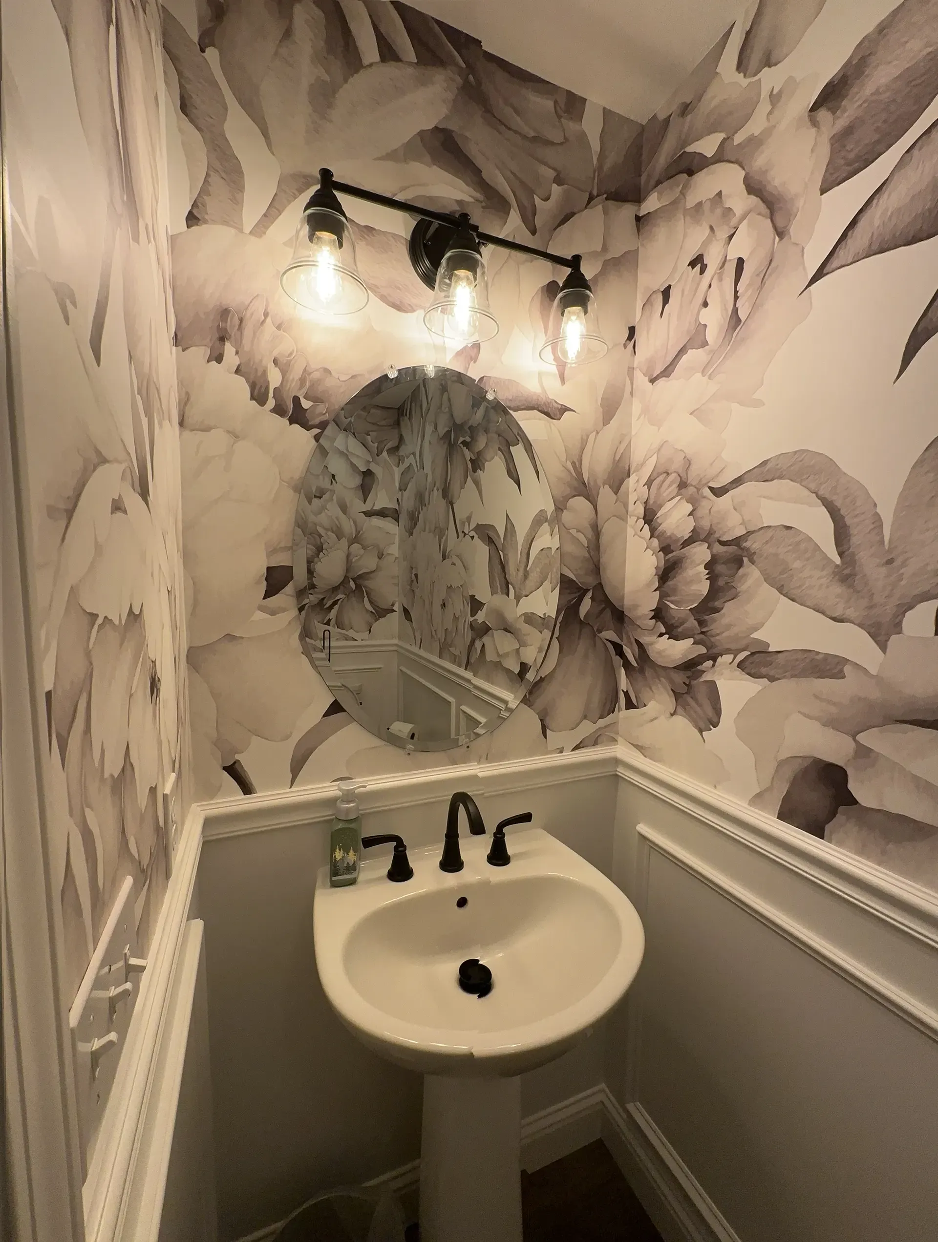 Small powder room with floral wallpaper, pedestal sink, and a round mirror.