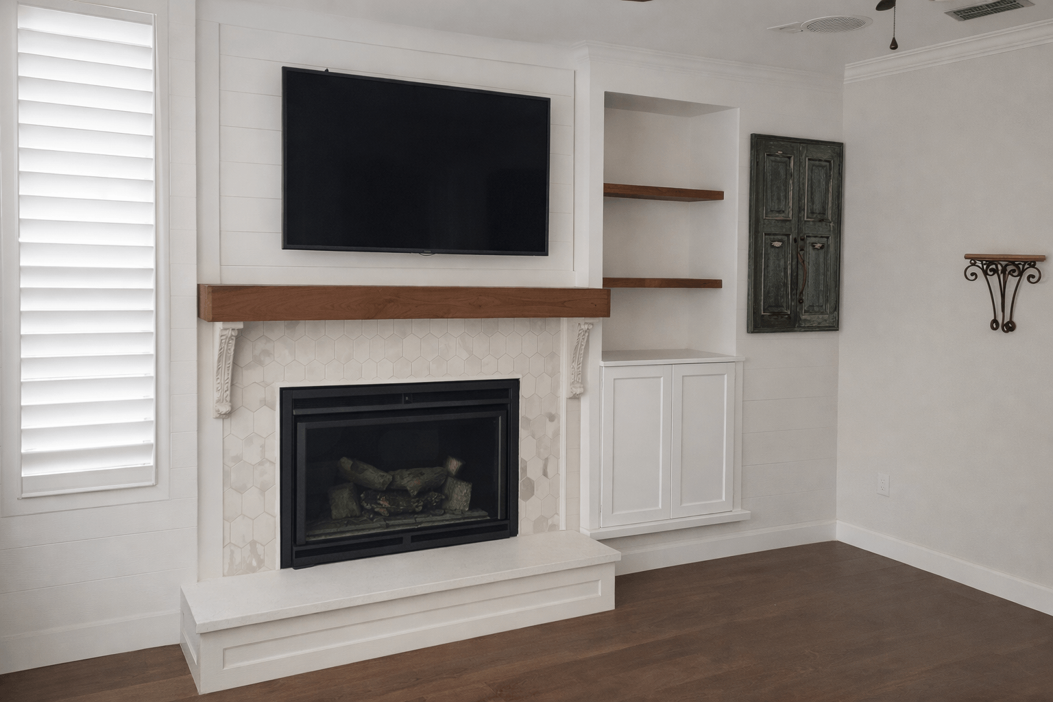 Living room with a fireplace, built-in shelves, TV, and shutters; light-colored walls and brown floor.