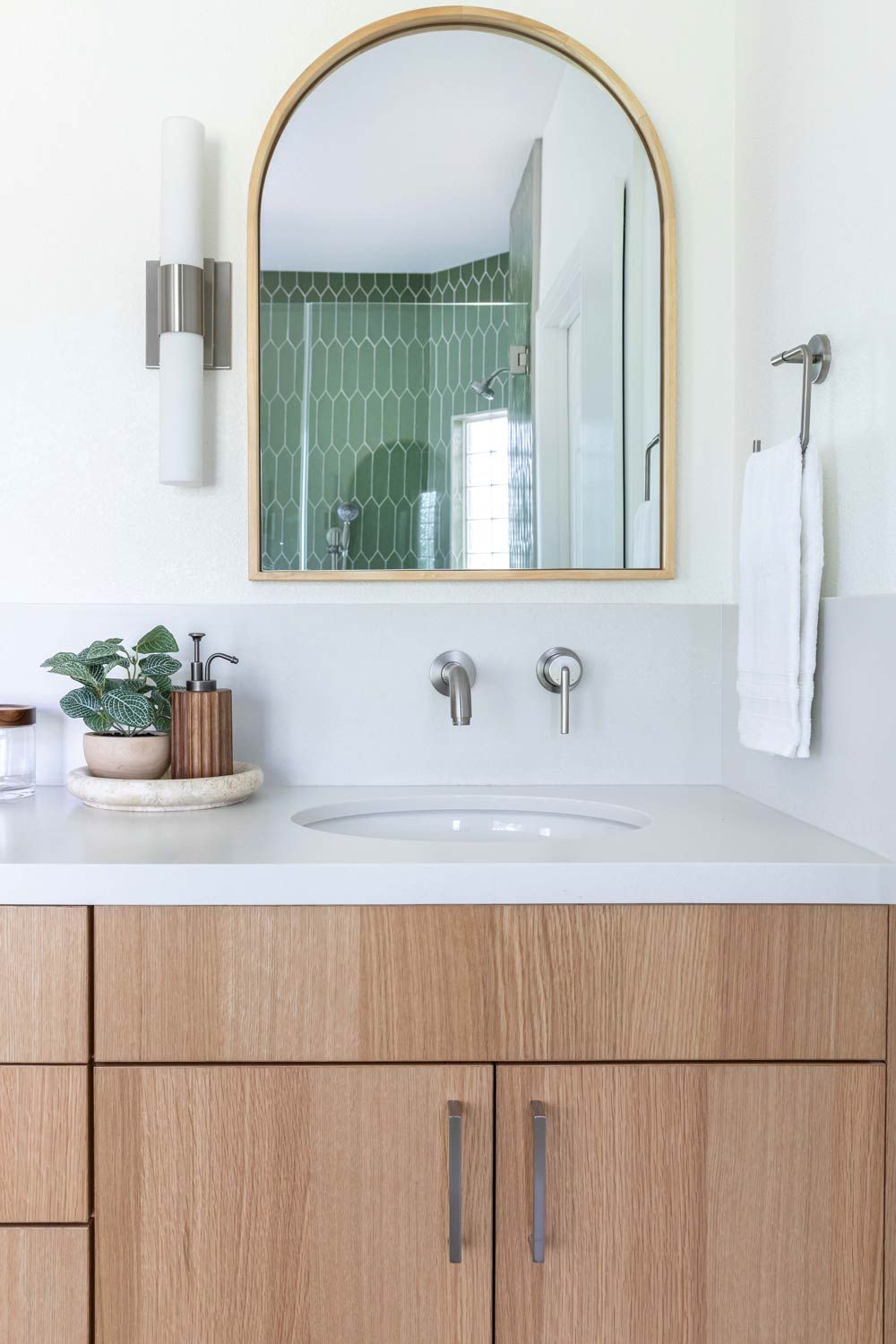Bathroom with a light wood vanity, arch mirror, and green tile in the background.