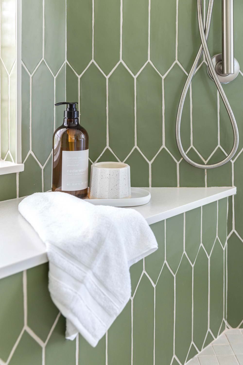 Green tiled shower with white shelf holding soap, cup, and towel. Silver shower head hangs to the right.