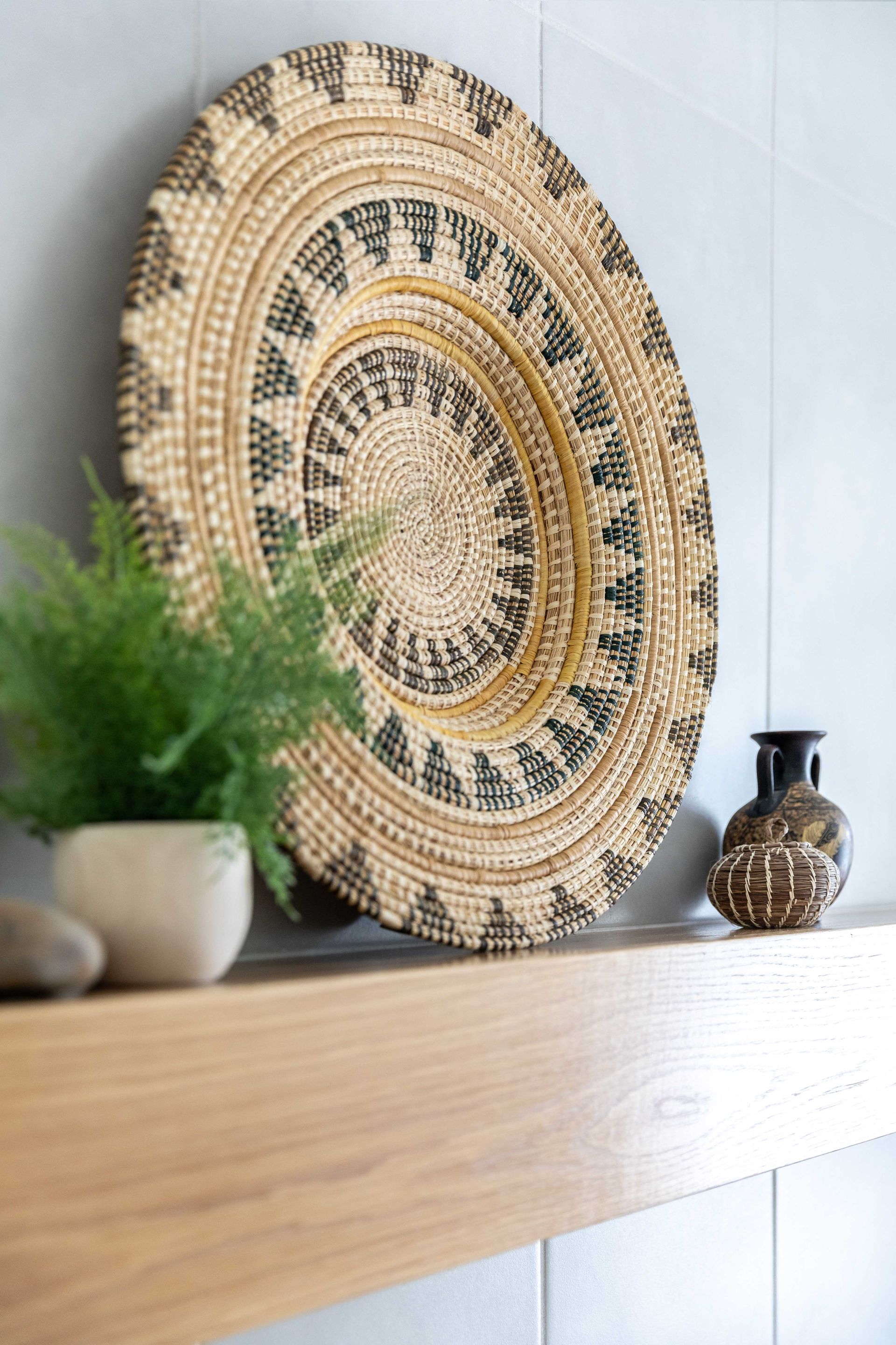 Woven decorative plate with a geometric pattern, on a wooden shelf, beside a fern in a pot and a small vase.