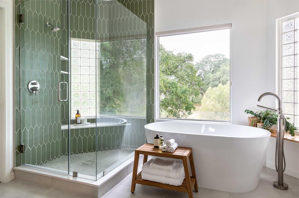 Modern bathroom with green tiled shower, white tub, and window overlooking trees.