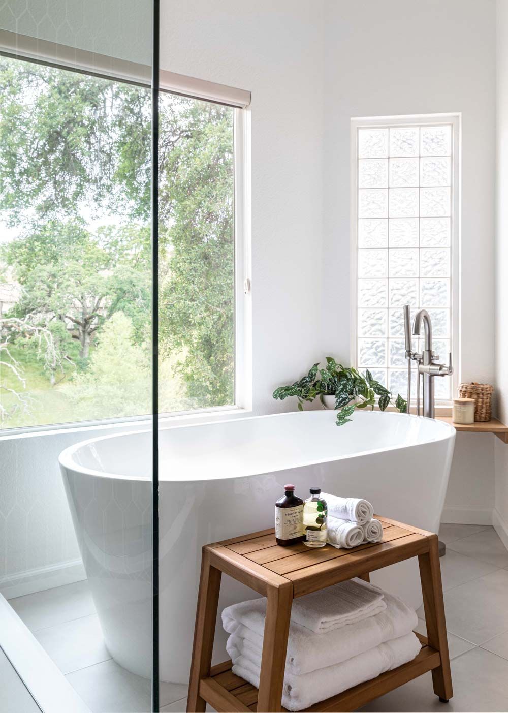 Bathroom with a white tub, wooden stool, large window with a view of trees, and glass block window.