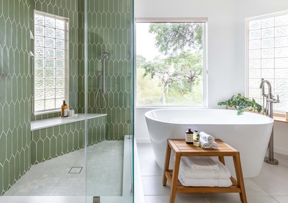 Green-tiled shower with glass door, adjacent to a white tub by a window. Wooden stool with towels sits beside the tub.