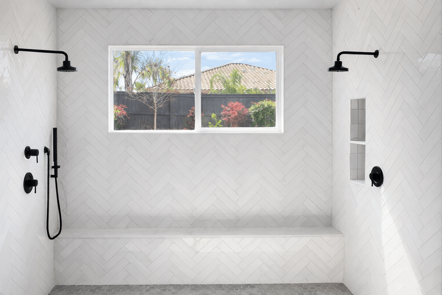 Modern white tile shower with black fixtures, bench, and window overlooking a yard.