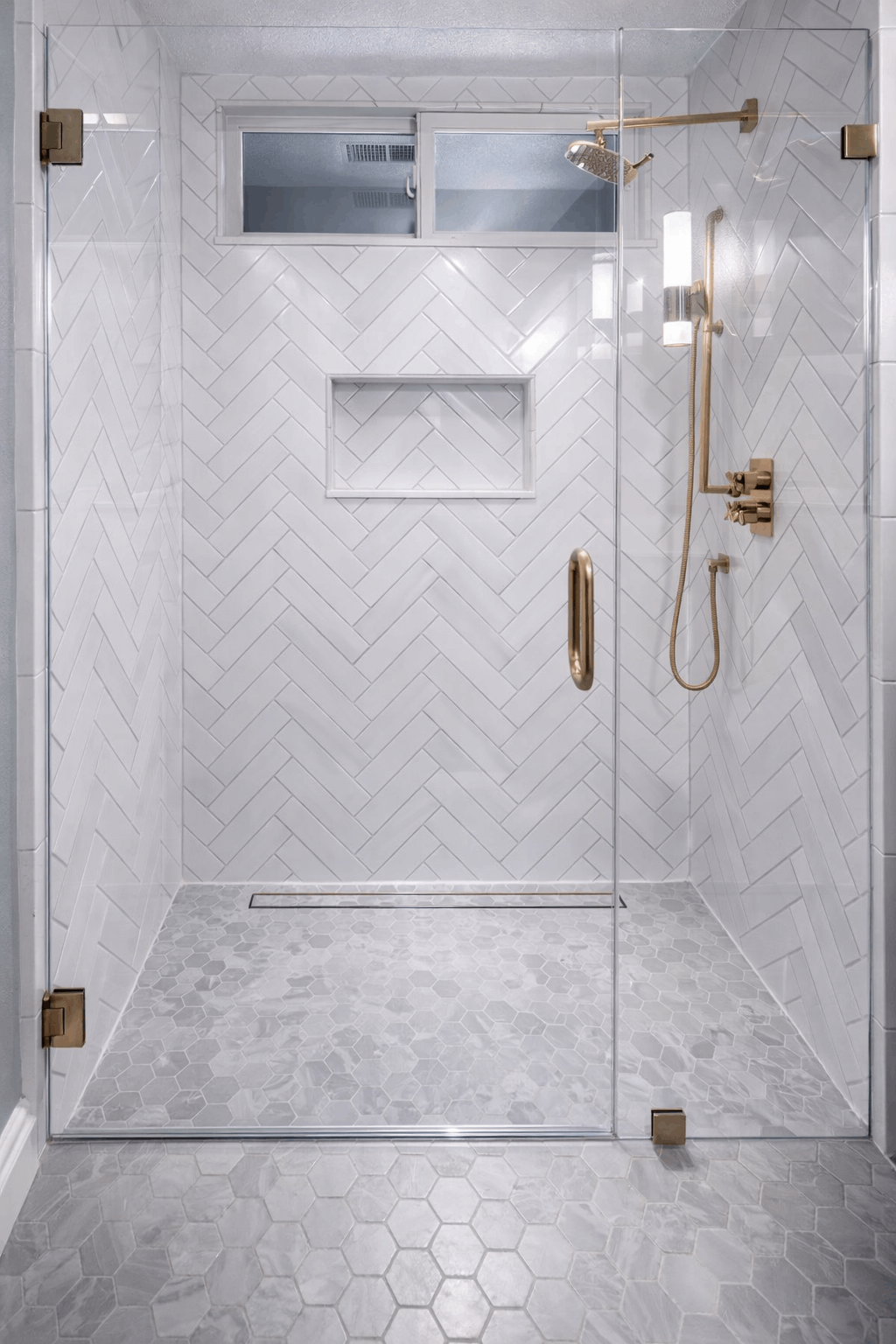 Glass-enclosed shower with white herringbone tile and gold fixtures. Hexagon floor tile. Recessed shelf.