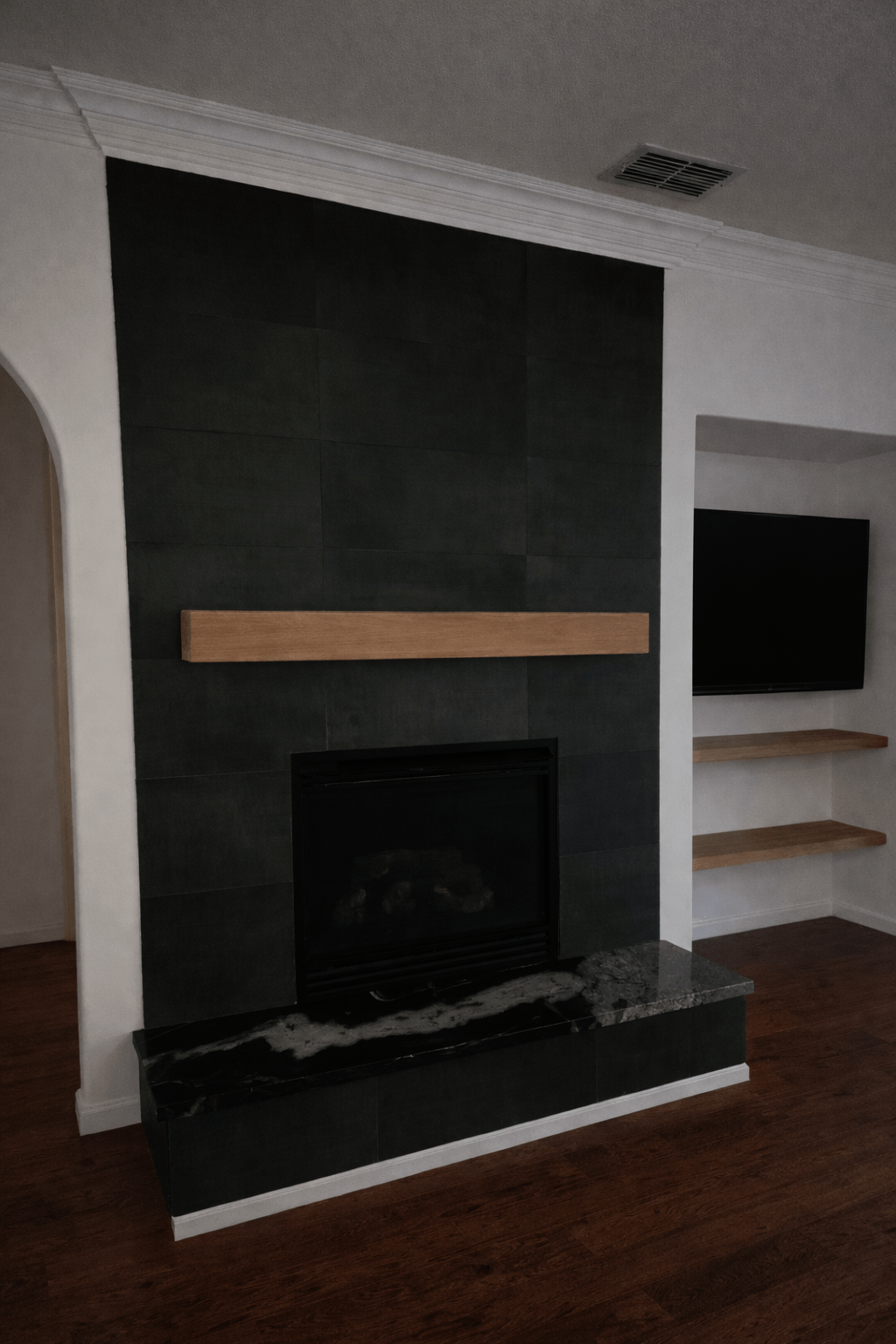 Black tiled fireplace with a wood mantel and matching shelf in an alcove with a wall-mounted TV.