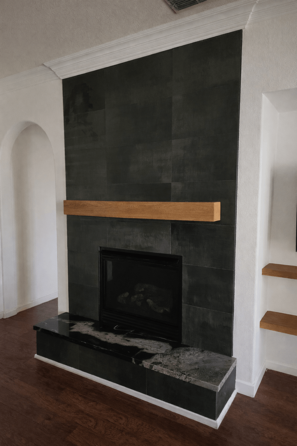 A black-tiled fireplace with a natural wood mantel and a dark stone hearth in a living room with white walls.