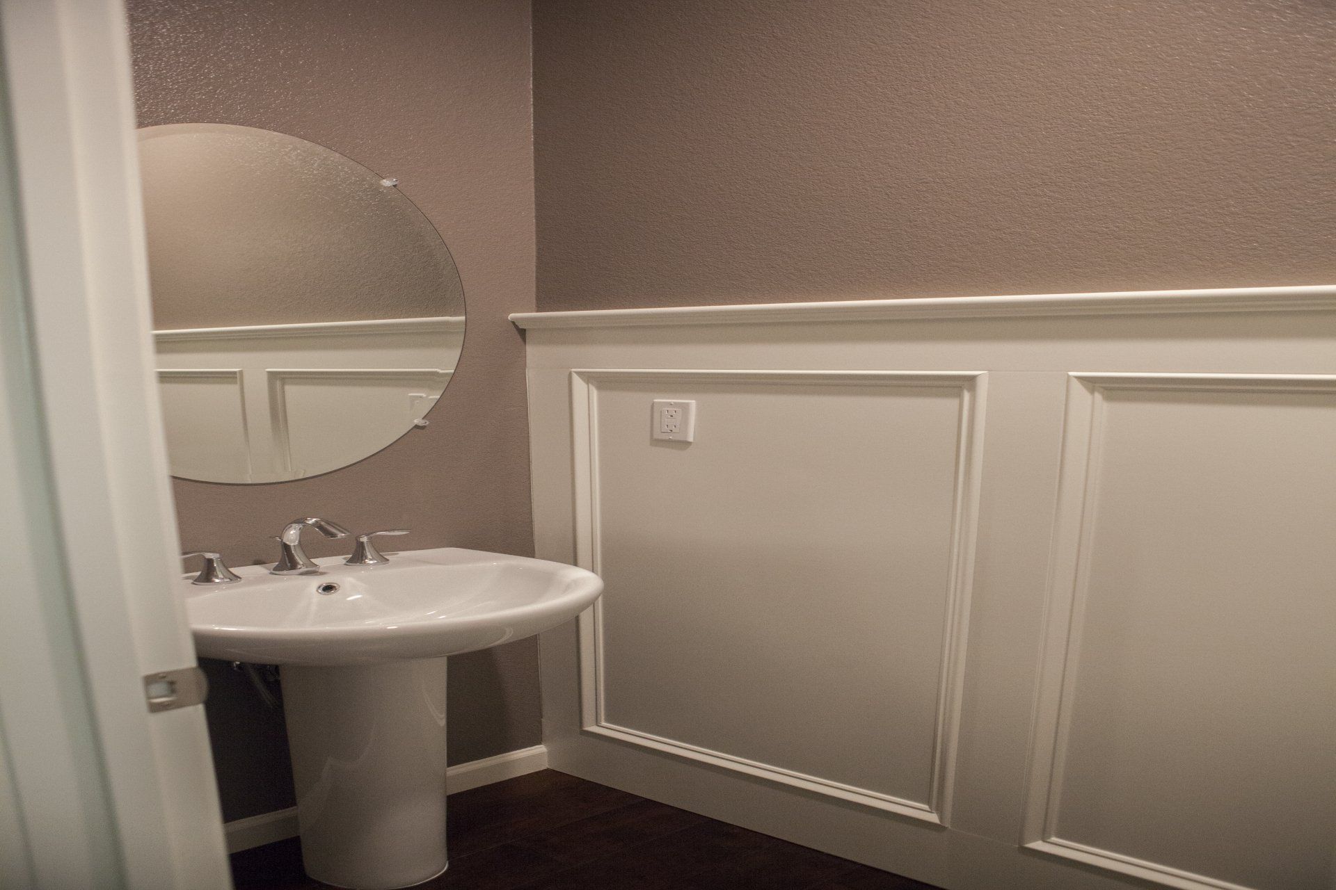 Bathroom with pedestal sink, round mirror, and textured gray walls. White wainscoting.