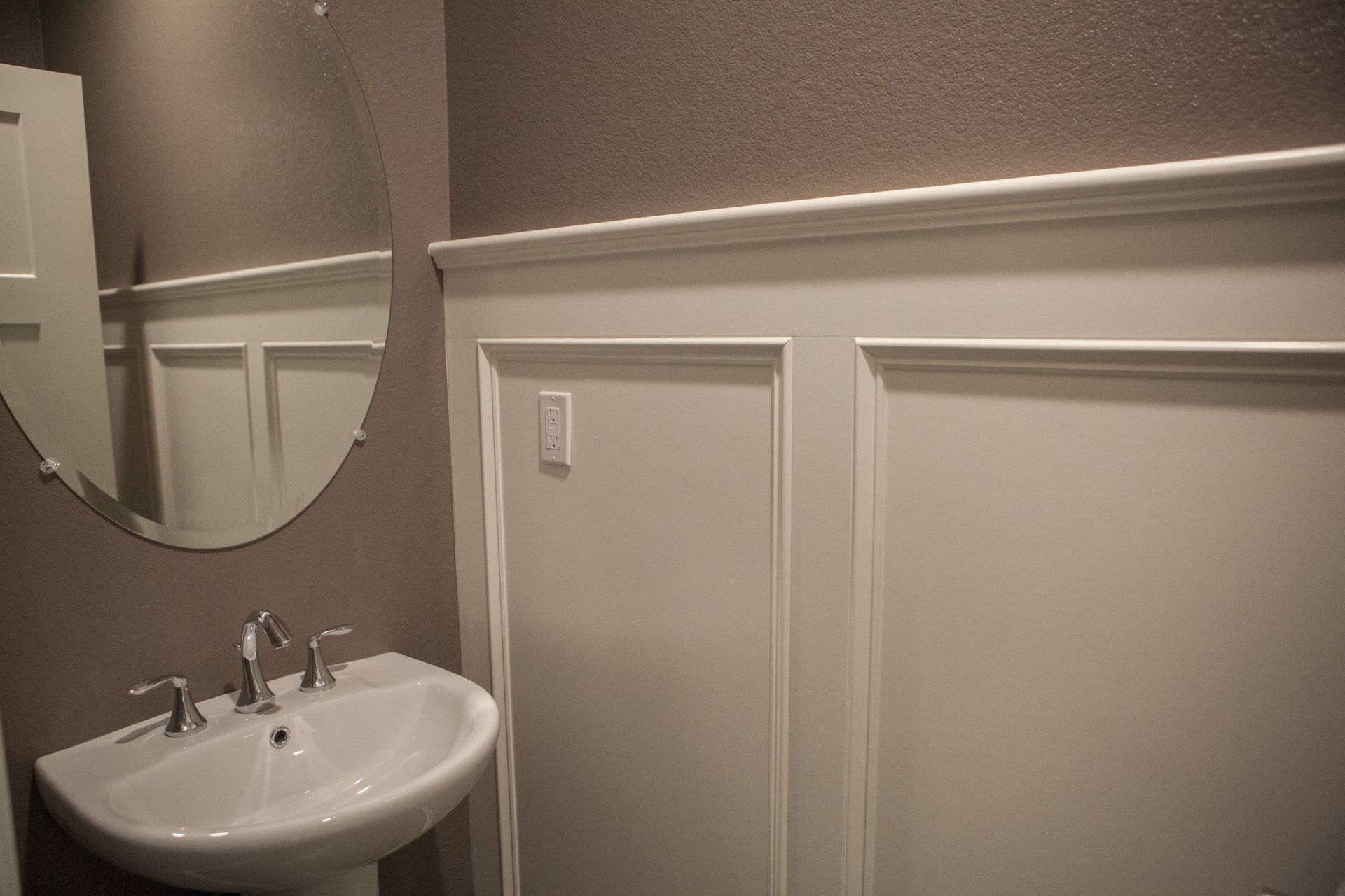 A bathroom with a white pedestal sink, round mirror, and white wainscoting.