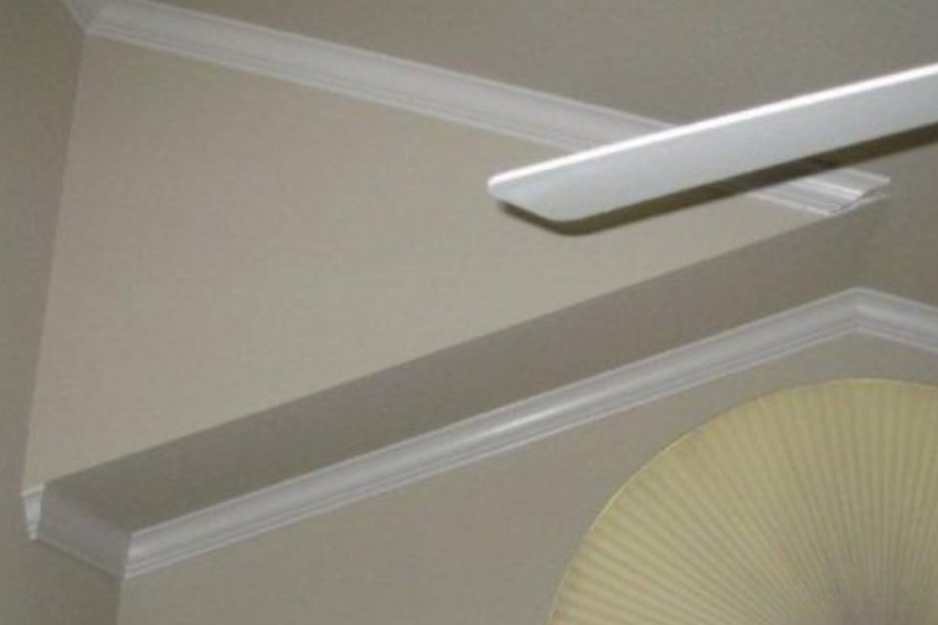 White crown molding on a tan wall, with a ceiling fan blade visible at the top and a partially visible fan at the bottom.