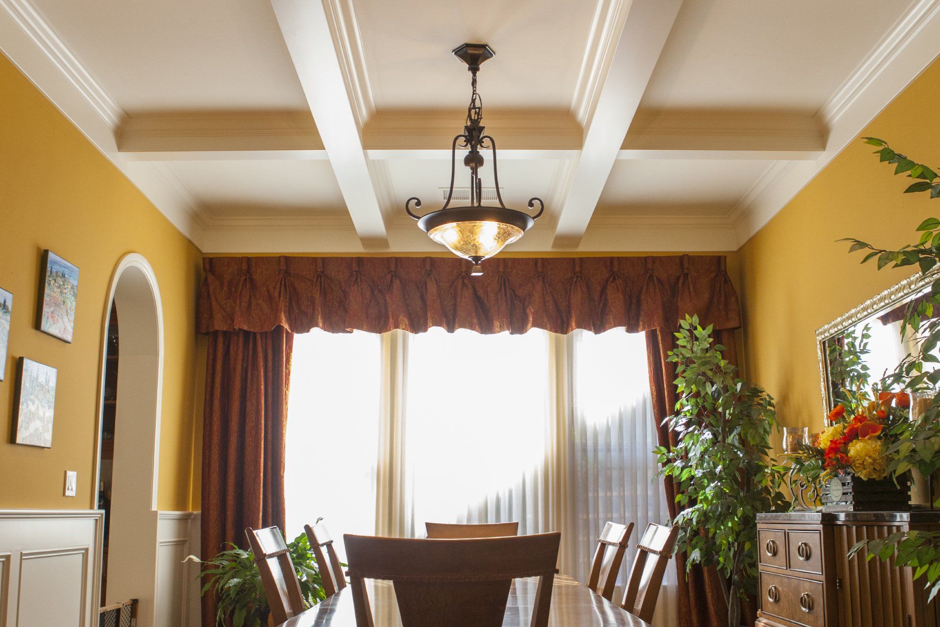 Dining room with a long table, chandelier, gold walls, and large window.