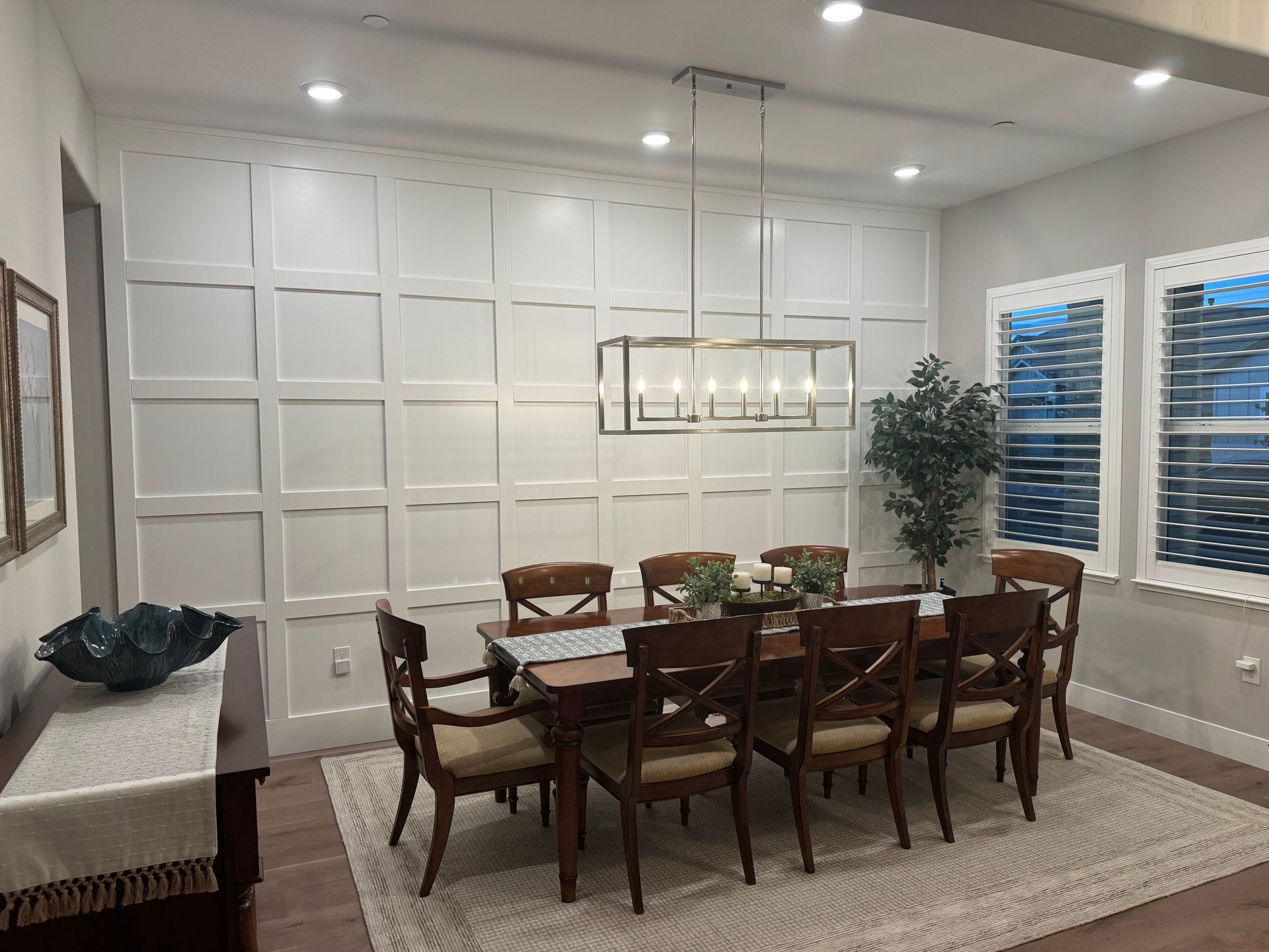 Elegant dining room with wooden table, chairs, rug, chandelier, paneled walls, and blue window blinds