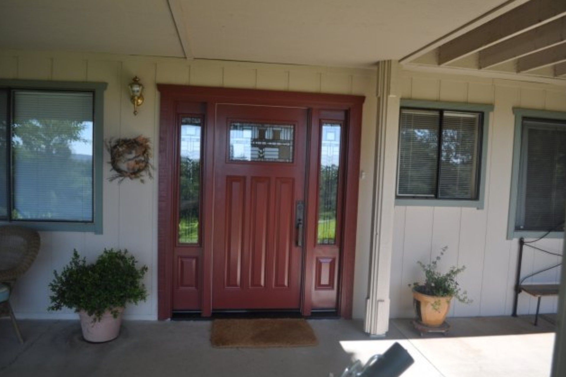 Red front door with sidelights, on a porch, flanked by windows and potted plants.