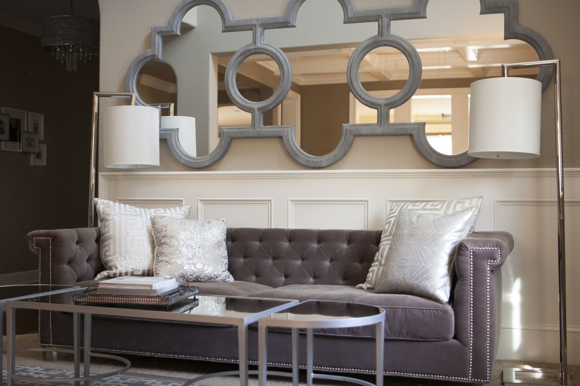 Gray tufted sofa with silver pillows, matching tables, large ornate mirror, and lamps.