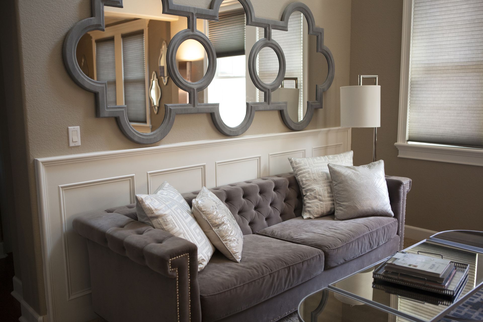 Gray tufted sofa with decorative pillows, beneath a large ornate mirror, against a beige wall.