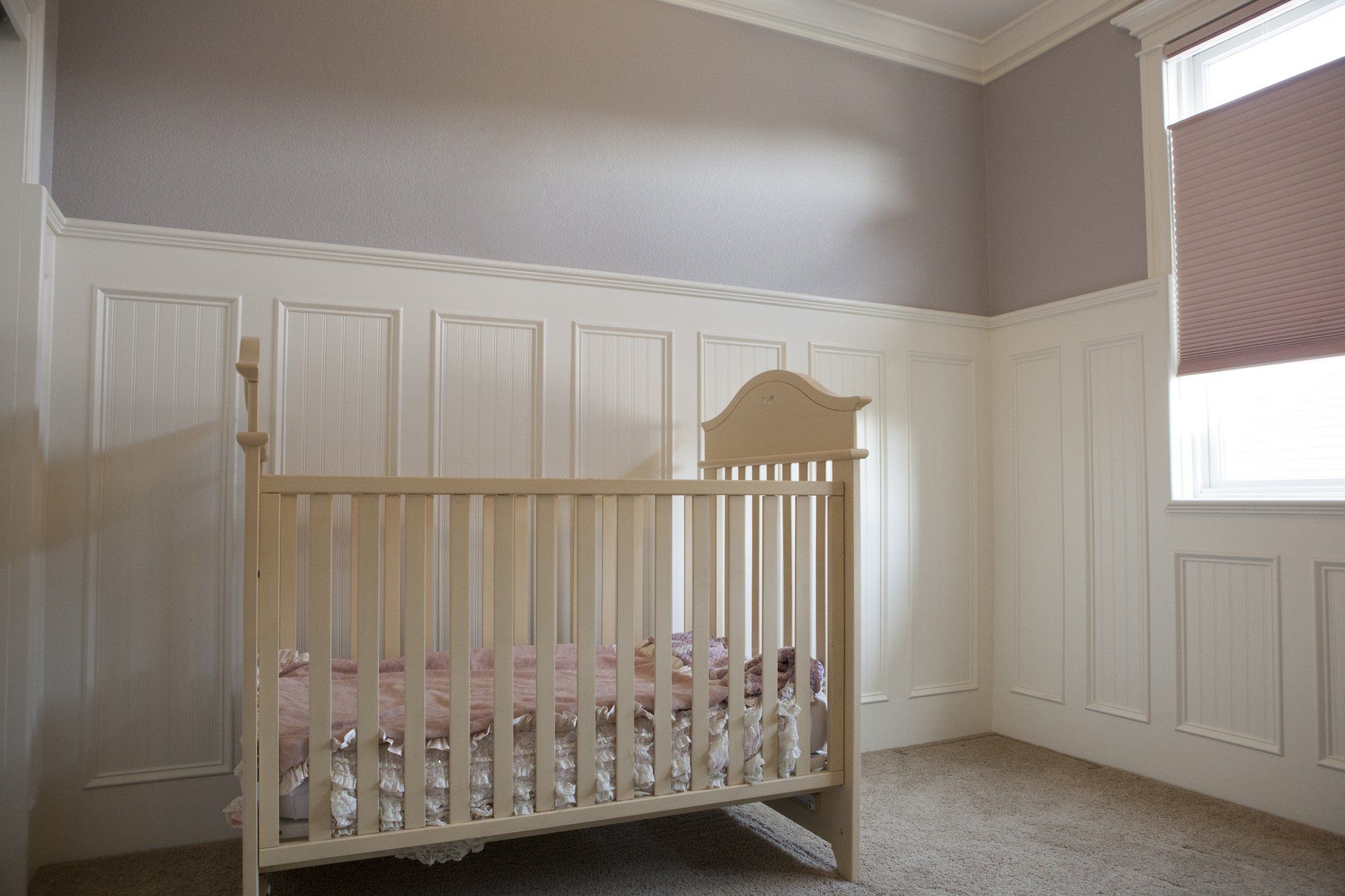 Crib in a nursery with white paneling and gray walls. Window with a pink shade.