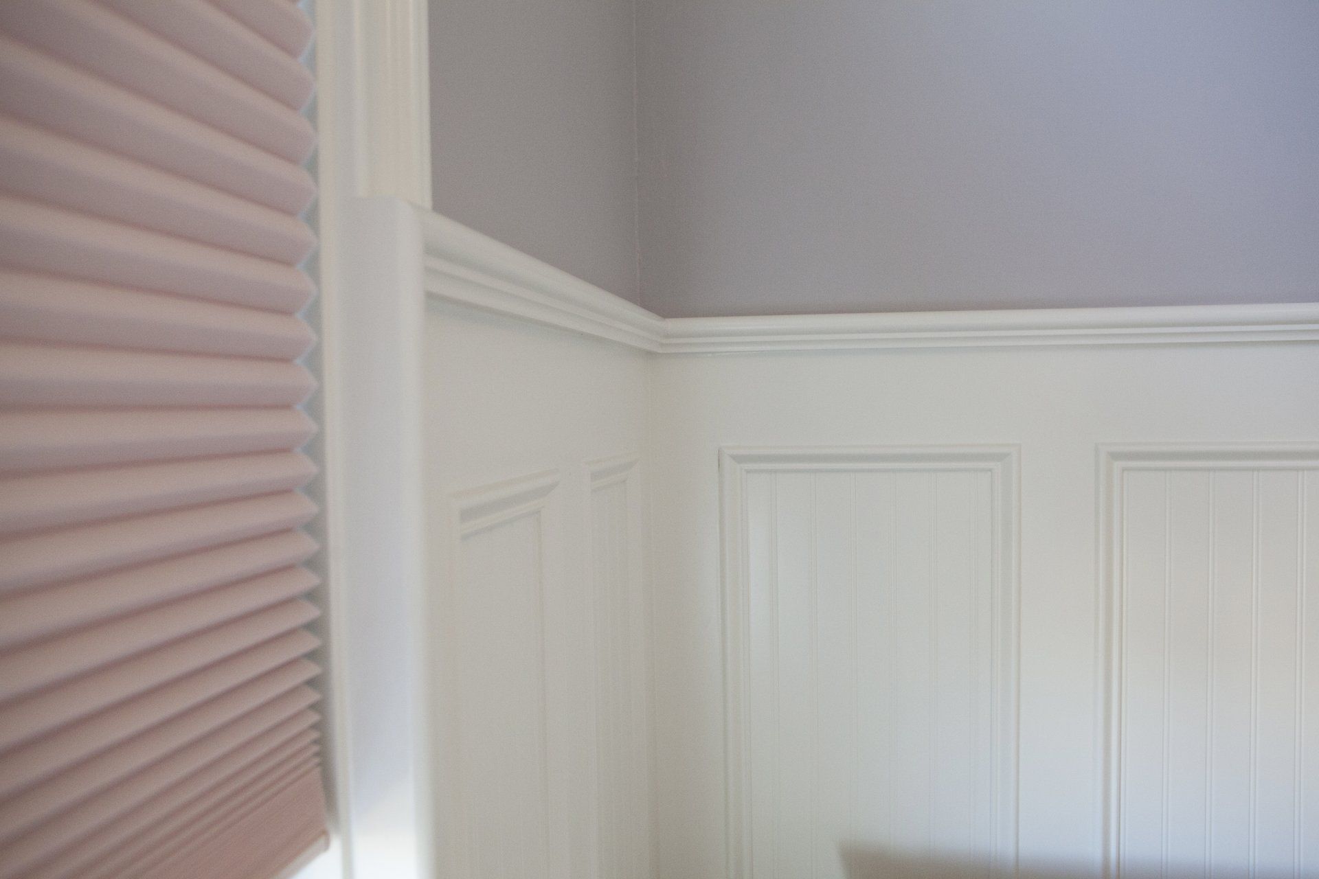 Corner of a room with white paneling, crown molding, and a purple wall above. Pink window blind on left.