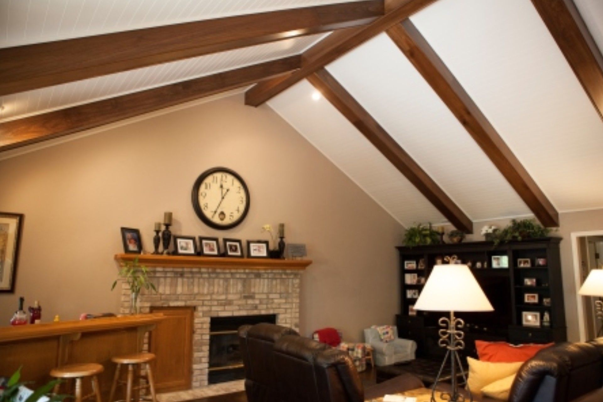 Living room with vaulted ceiling, fireplace, and dark wood beams.  Beige walls, brown trim, and large clock.