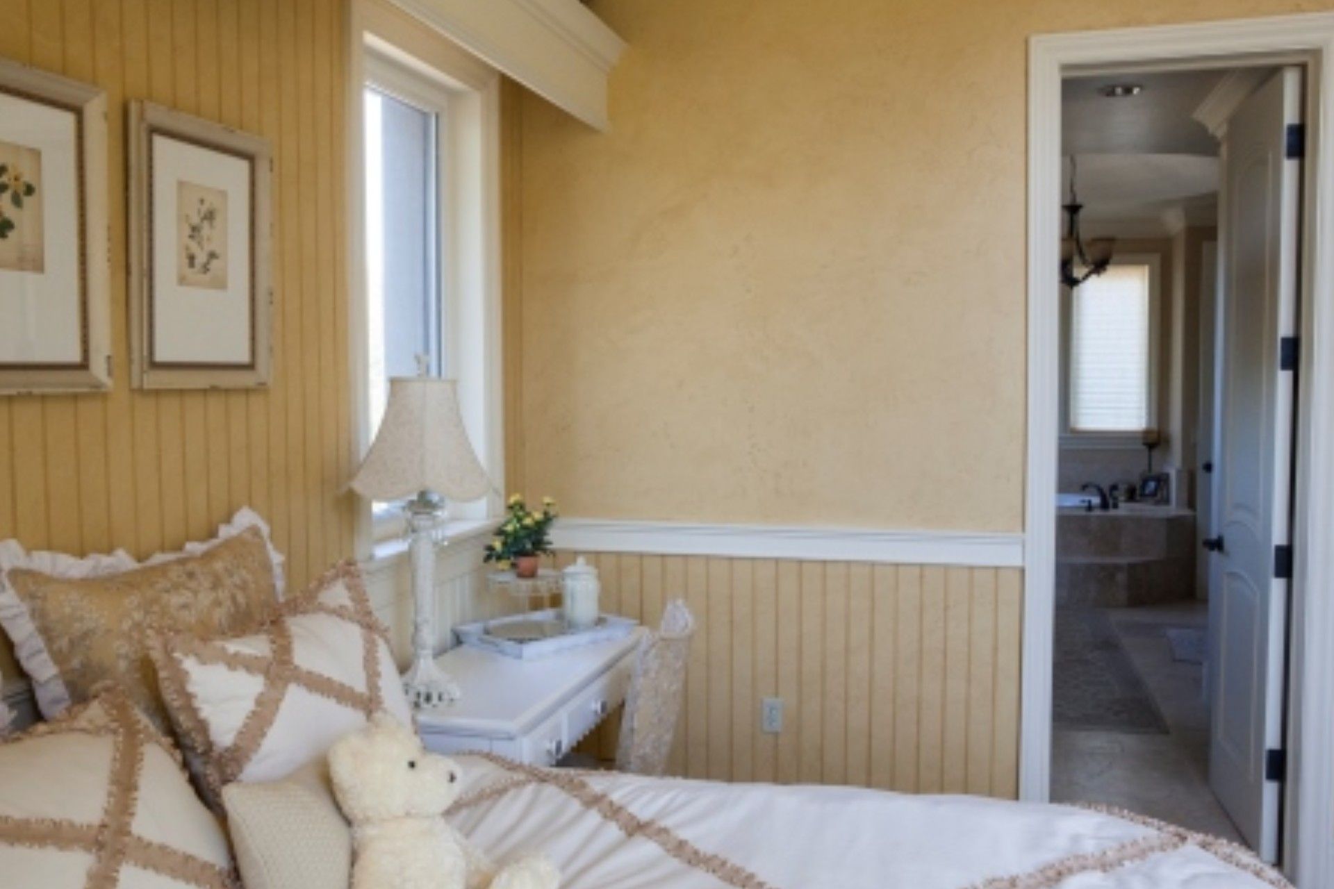 Bedroom with yellow walls, white trim, bed with pillows and teddy bear, door to bathroom.