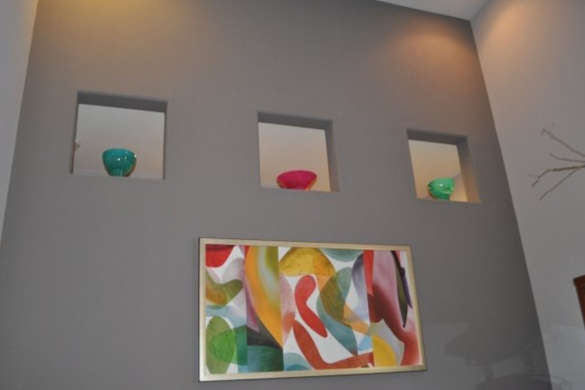 Gray wall with three recessed shelves holding colorful bowls; below, an abstract painting.