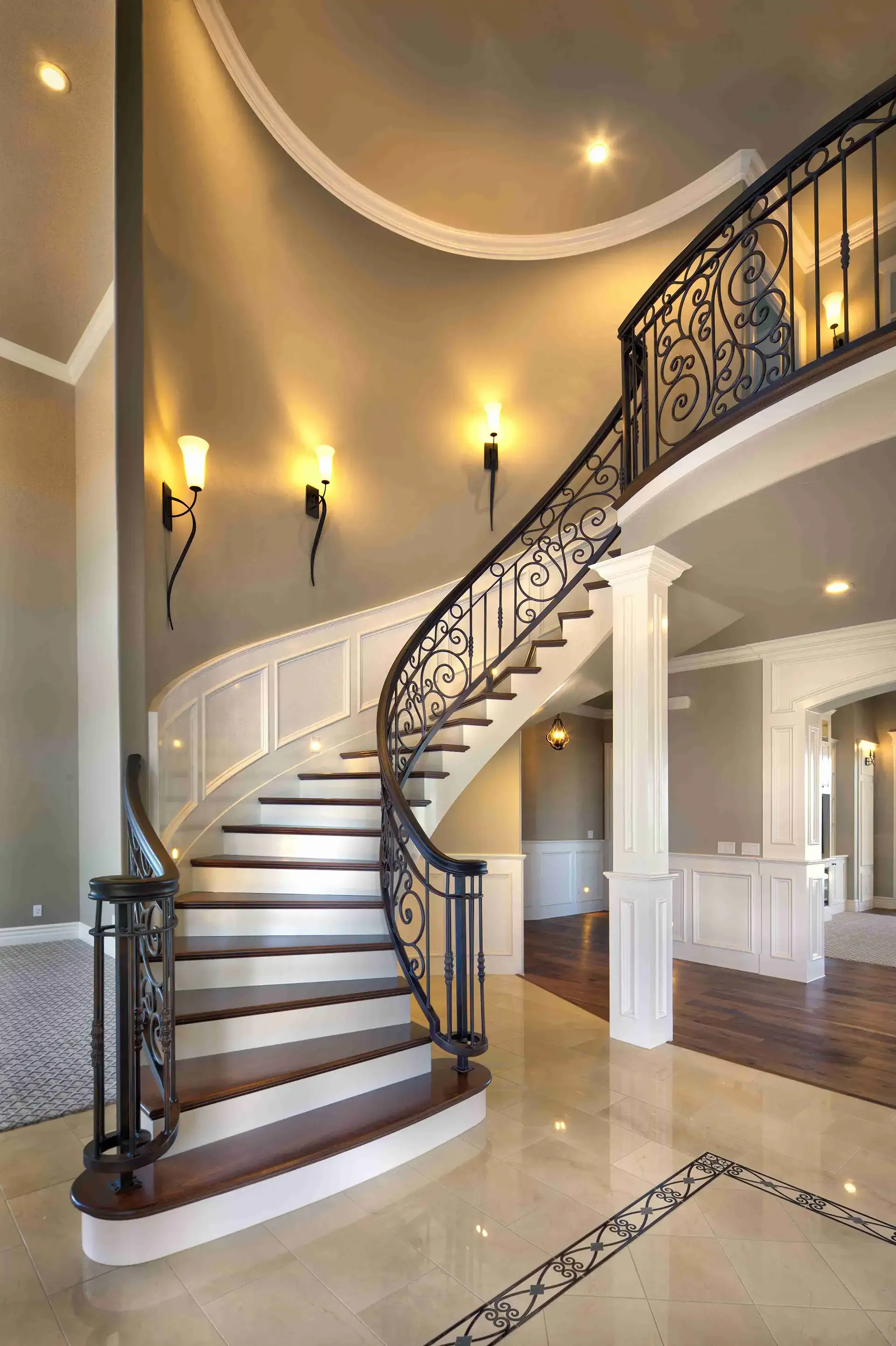Elegant curved staircase with ornate iron railing and sconce lighting in a grand foyer.