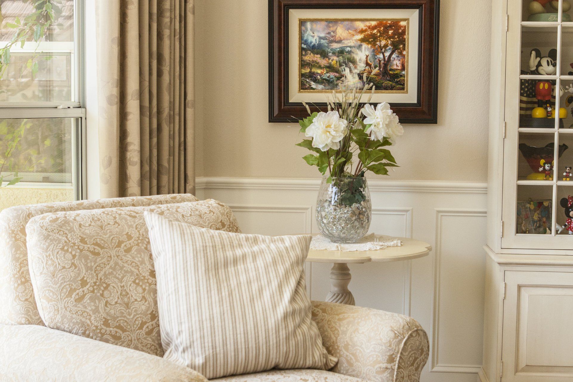 Cozy living space with beige sofa, white flowers on a side table, painting on wall, and a cabinet.