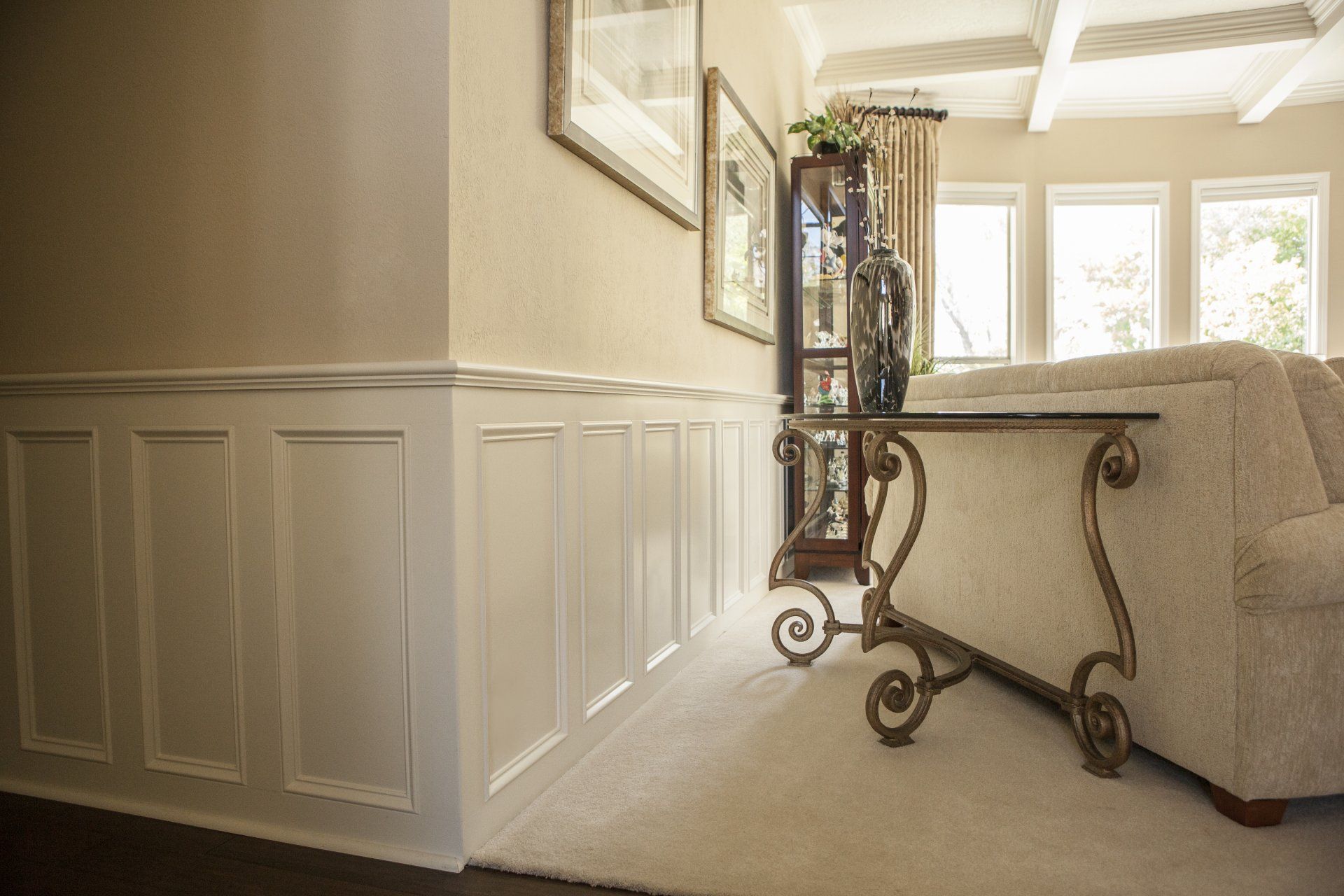Beige living room with white wainscoting, a decorative table, and a cream-colored sofa near windows.