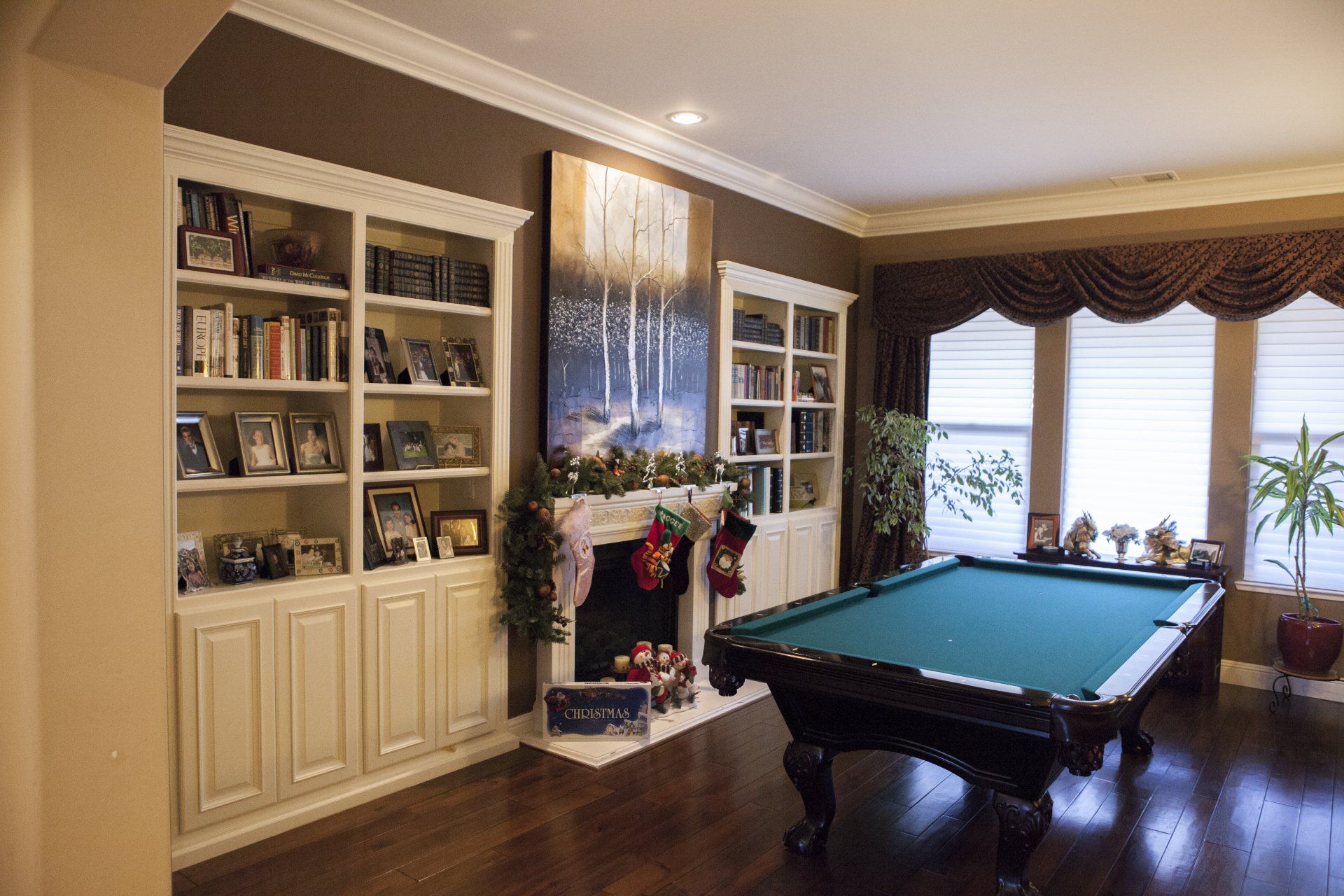 Pool table in a room with built-in bookshelves, fireplace, and large windows with brown curtains.