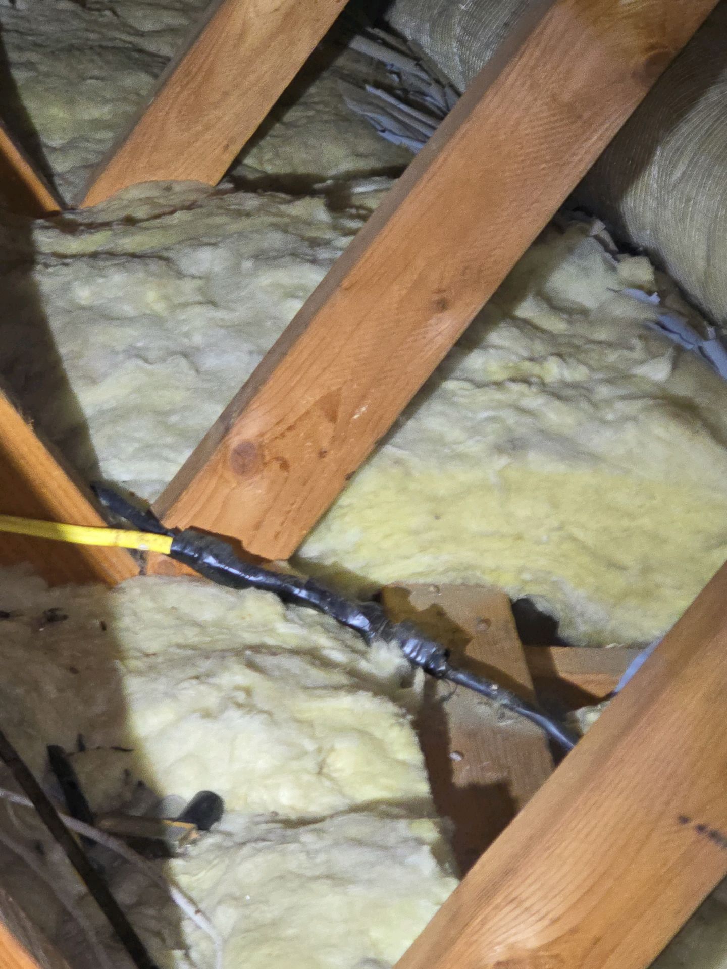 Electrical wires joined with black electrical tape resting on yellow fiberglass insulation between wooden attic rafters.