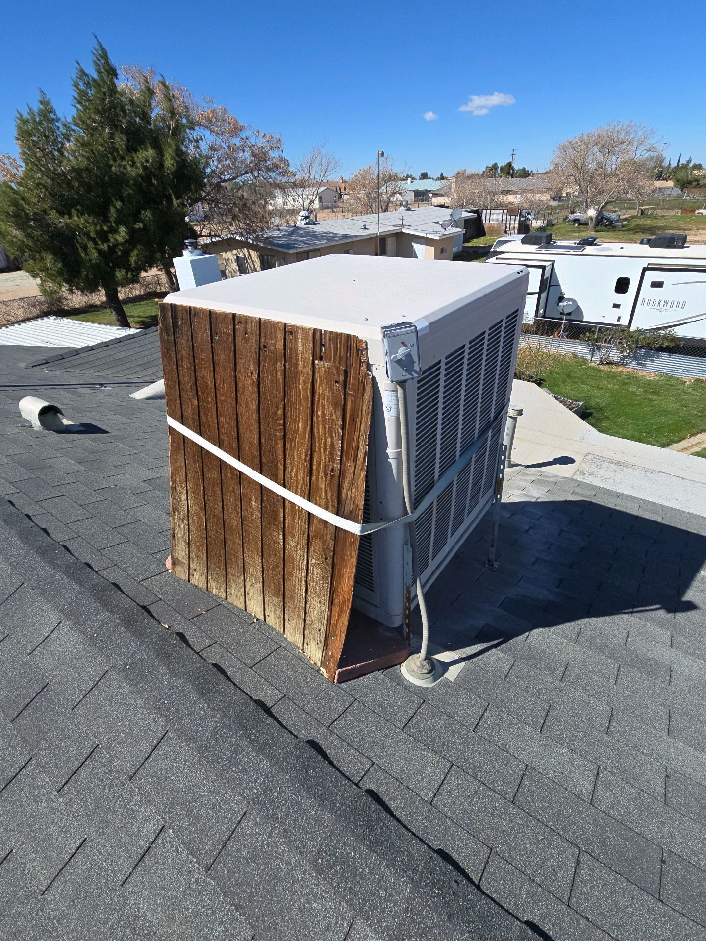 A residential rooftop evaporative cooler with a partially detached, weathered wooden side panel secured by a white strap.