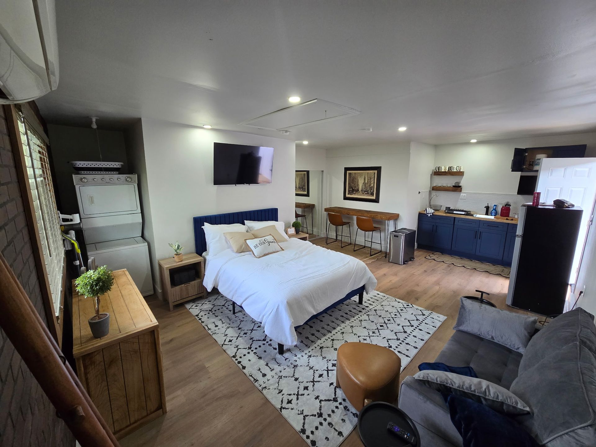 A bright studio apartment with a bed, a gray sofa, a wooden dresser, and a small kitchen area with blue cabinets.