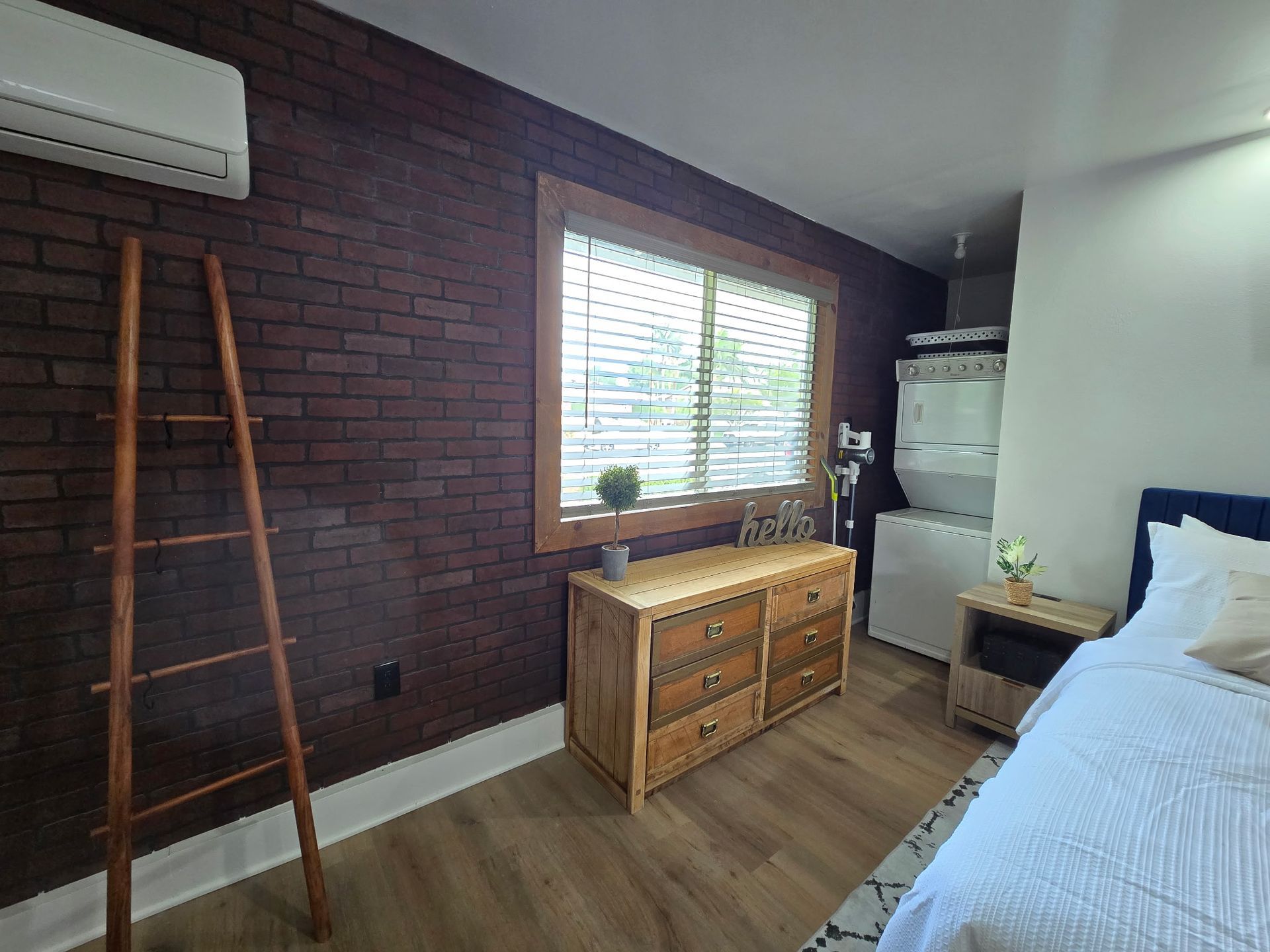 A bedroom with a dark brick wall, wooden ladder, dresser, window, stacked laundry unit, and part of a white bed.