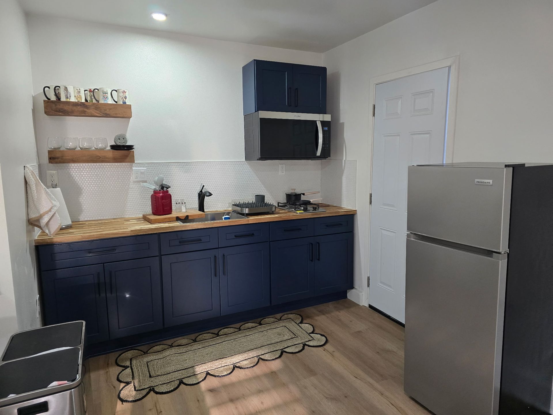 A small kitchen with navy blue lower cabinets, wood countertops, floating shelves, a microwave, and a stainless fridge.