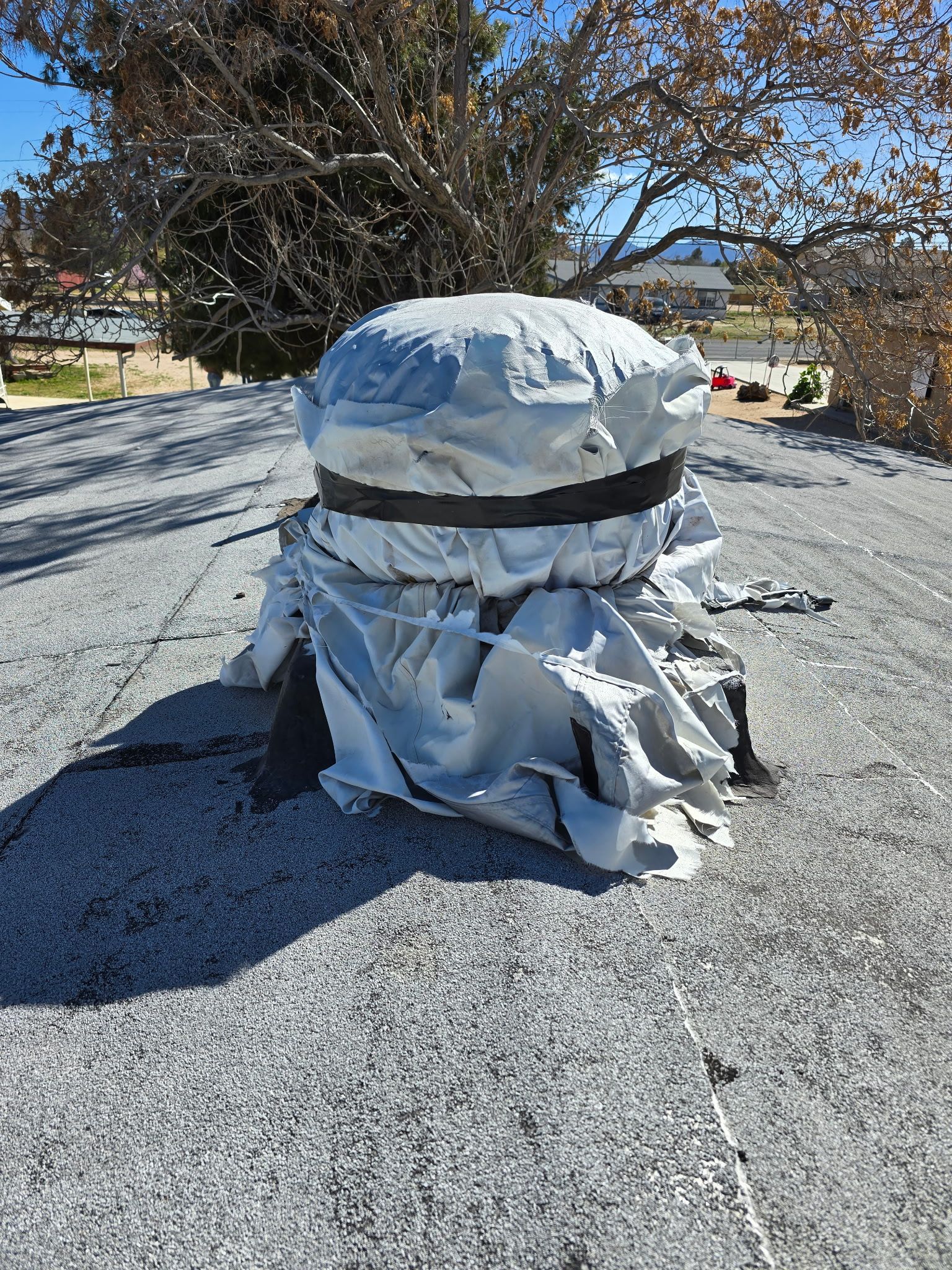 A damaged, silver-colored ventilation cover wrapped in a dark strap, sitting on a weathered, gray flat roof.