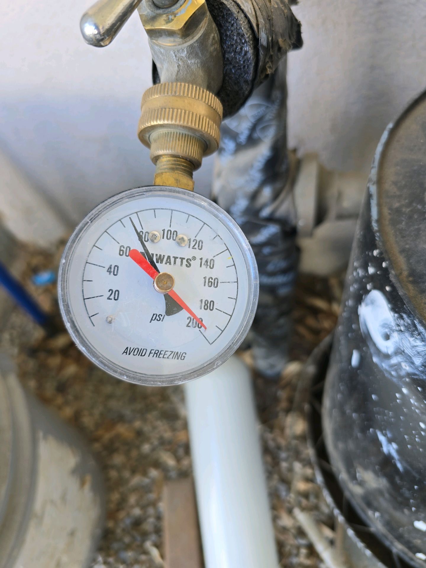 A circular analog pressure gauge attached to a metal faucet, showing the red needle pointing just below 60 PSI.