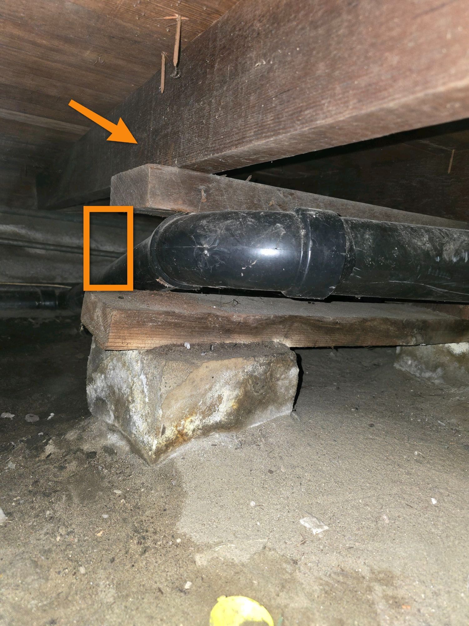 Crawl space view showing a black pipe resting on wooden blocks beneath wooden floor joists, with arrows highlighting it.