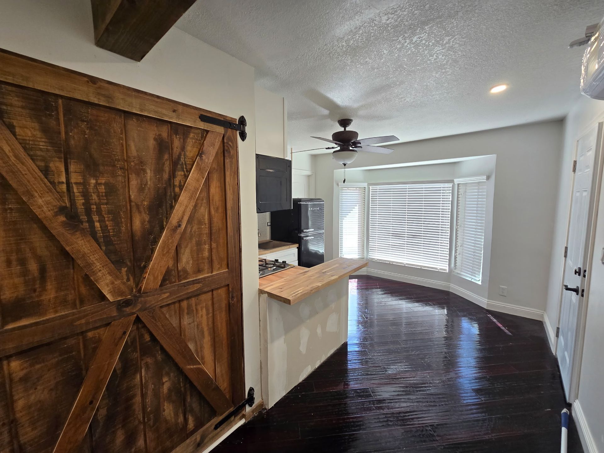 A room features dark wood flooring, a rustic barn door, a kitchen counter, and a ceiling fan in front of a bay window.