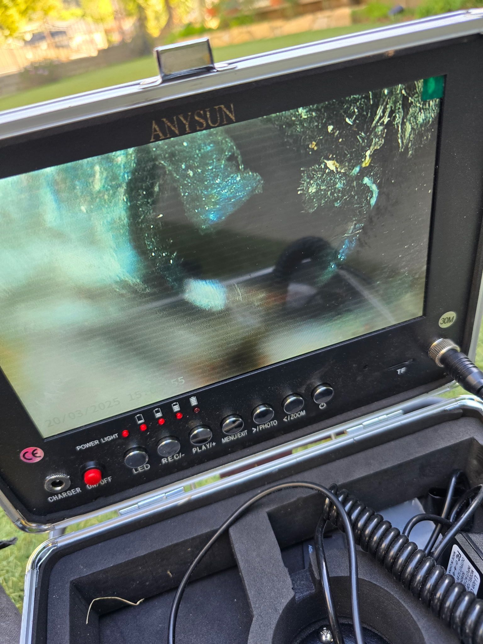 An Anysun sewer inspection camera monitor displaying a blurry, dark view of an underground pipe on its screen.