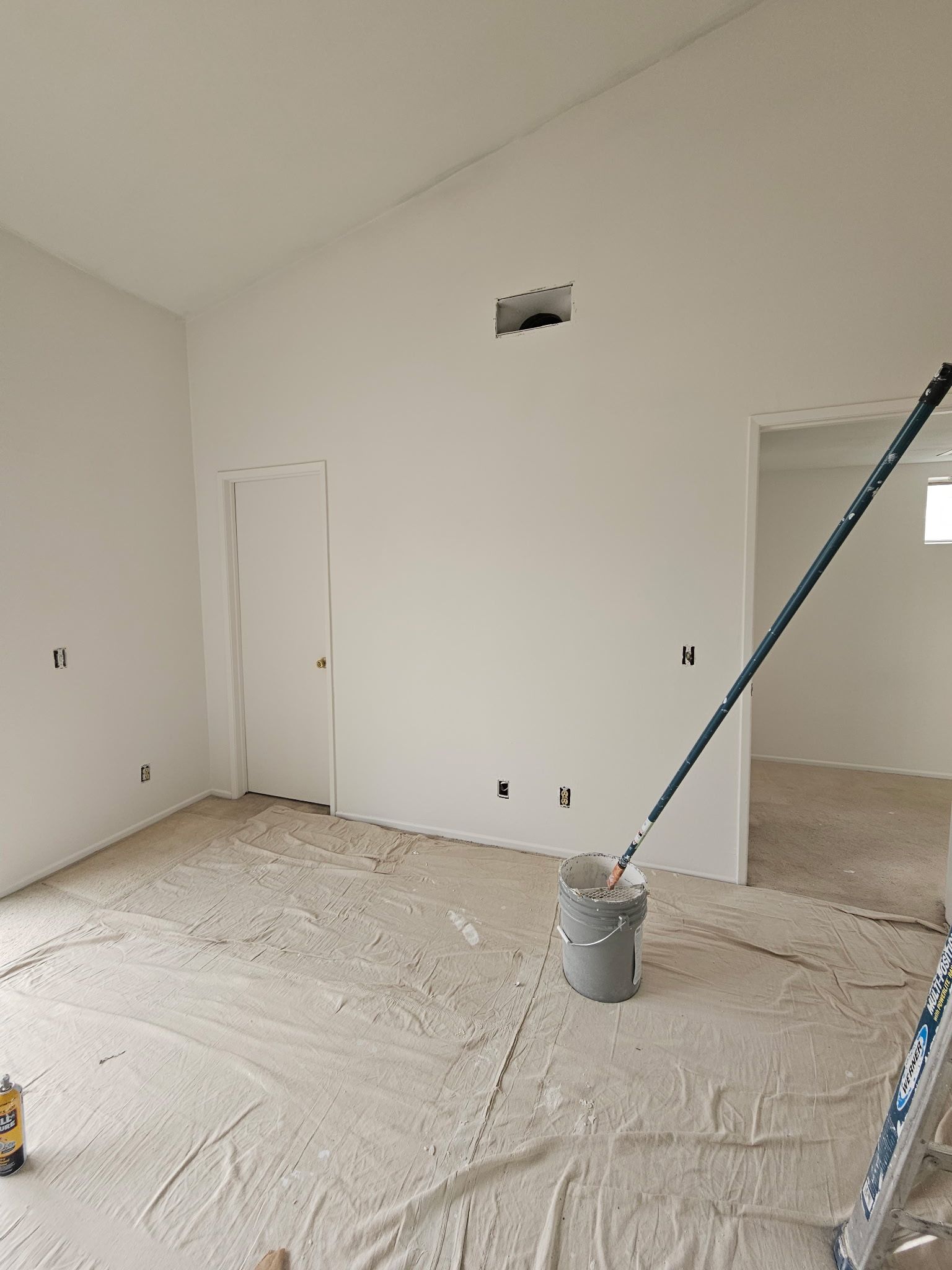 A white room under renovation with drop cloths, a paint bucket, a long-handled roller, and an open doorway.