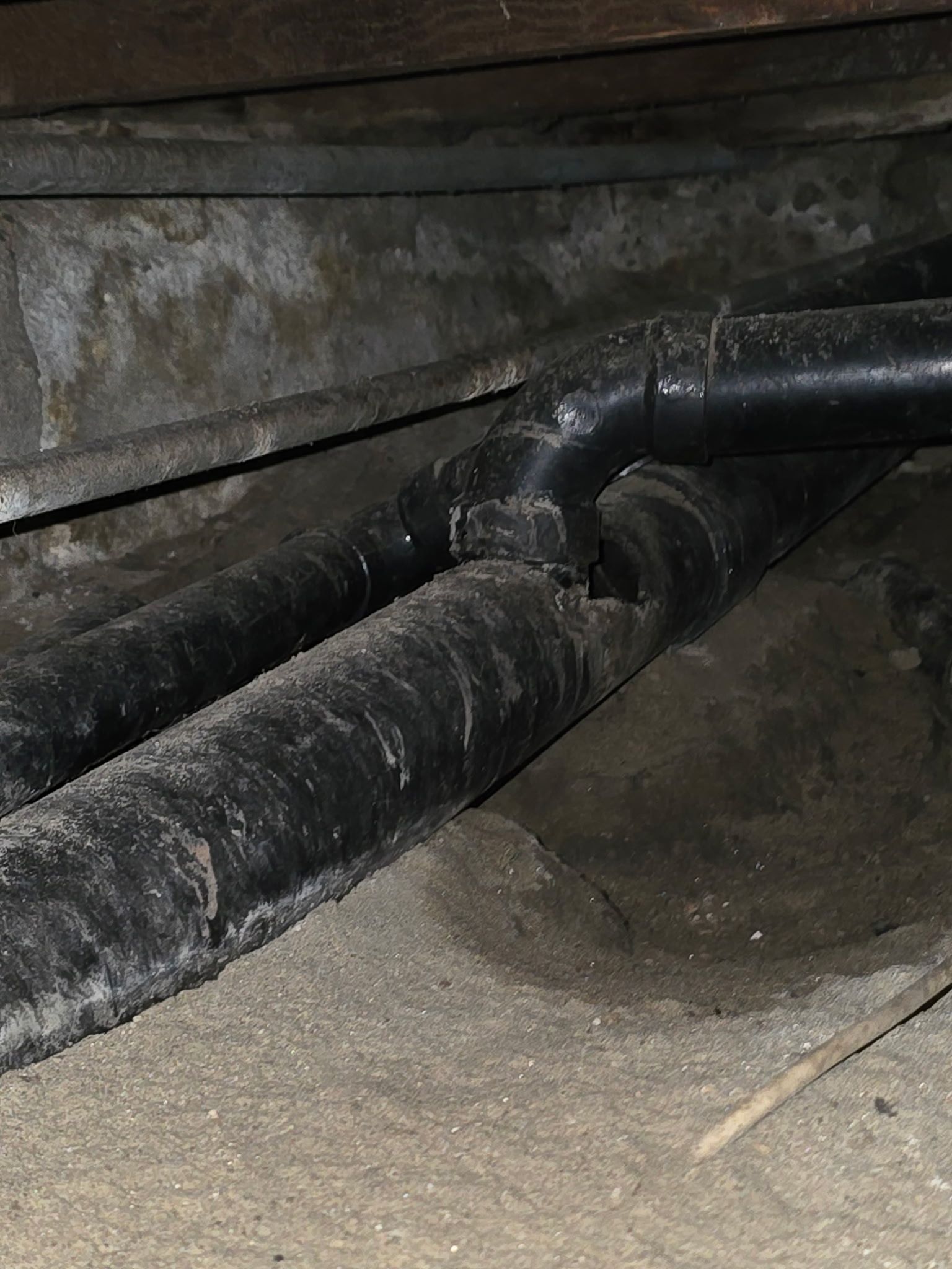 Black ABS plumbing pipes in a crawlspace, showing a junction where one pipe connects to a main horizontal line.