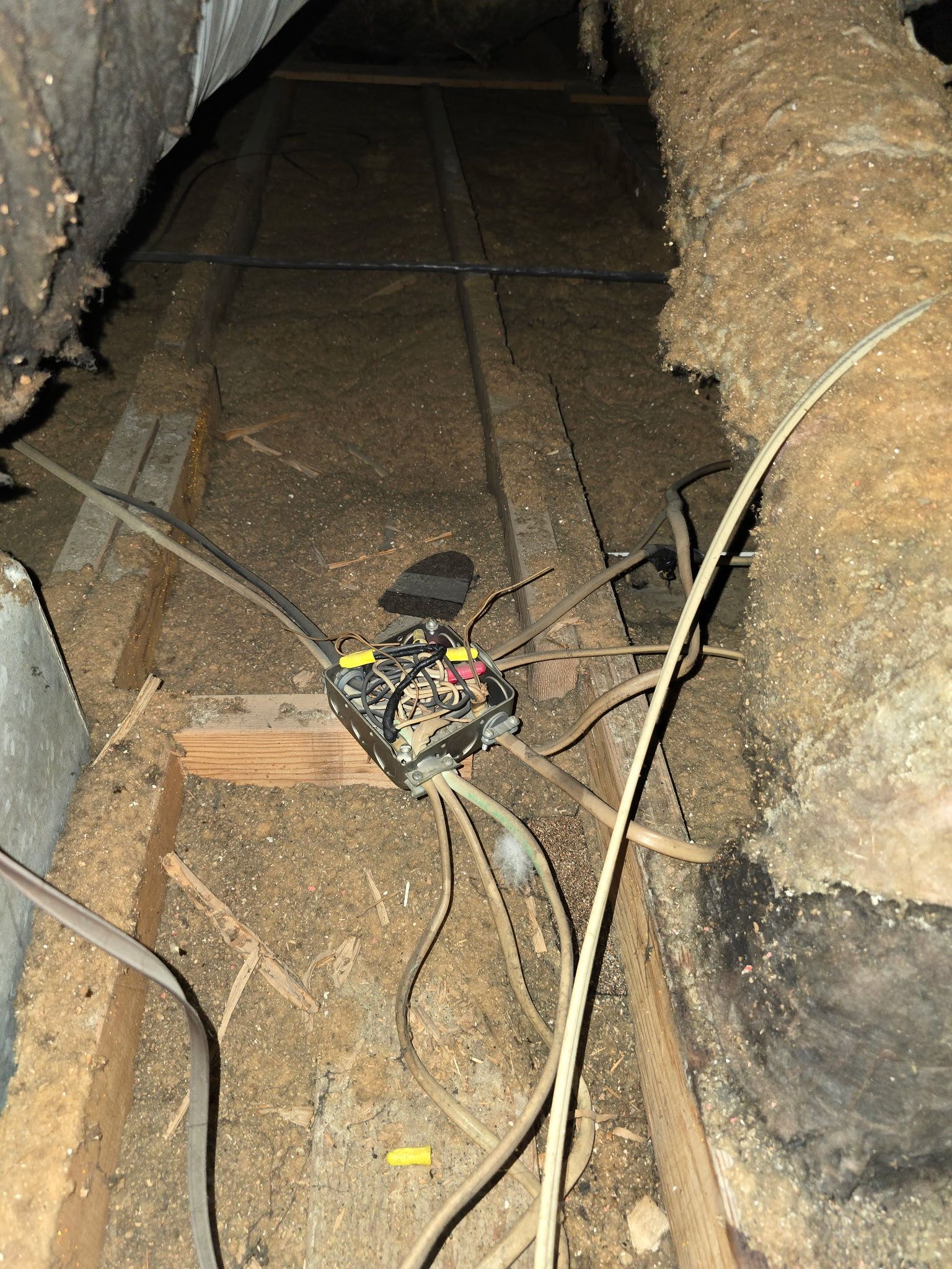 An electrical junction box with several wires lies exposed on a wooden beam in a dusty, dimly lit attic space.