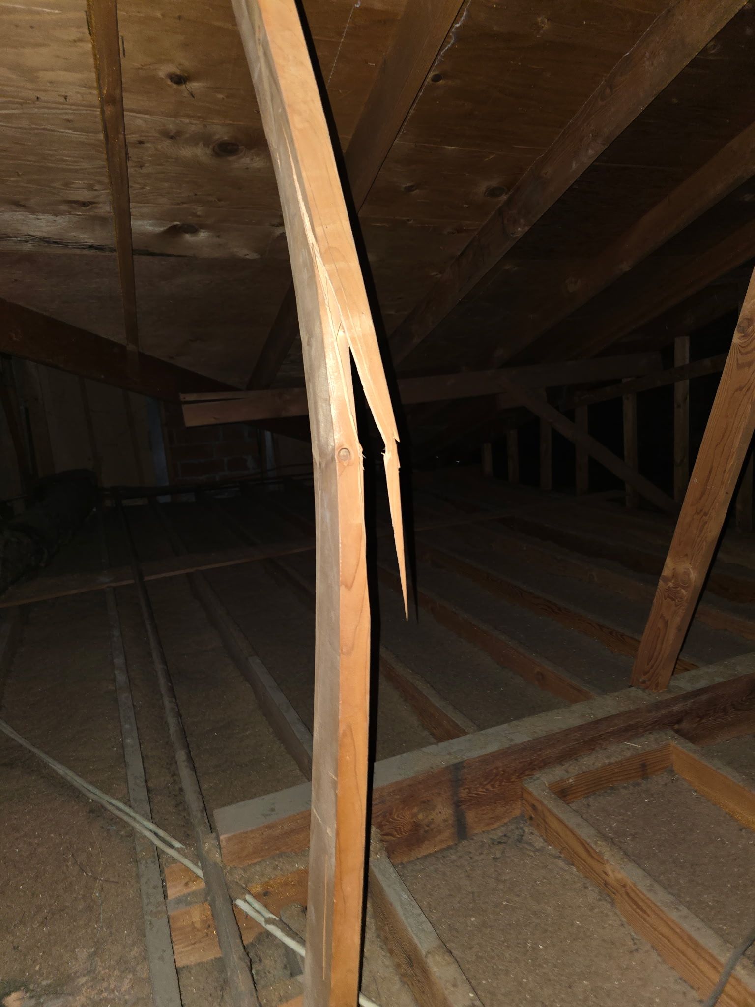 A split wooden support beam in a dark attic, showing structural damage to the vertical timber.