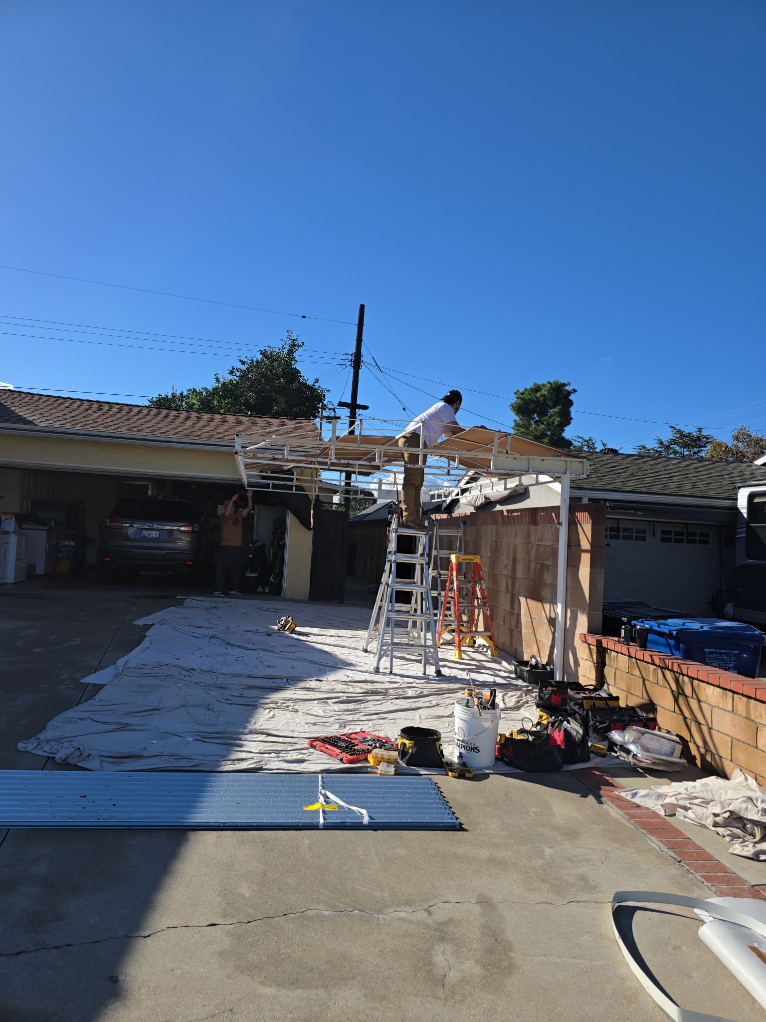 A worker stands on a ladder under a partially demolished white patio cover on a sunny day in a driveway.