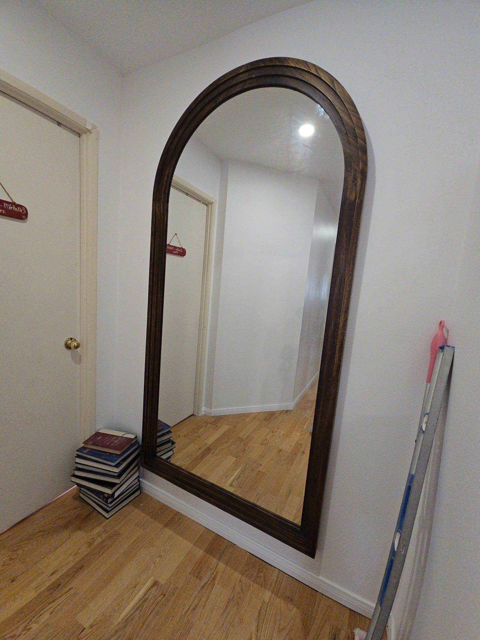 Full-length arched mirror on a white wall in a hallway with wood floors and nearby stacked books.