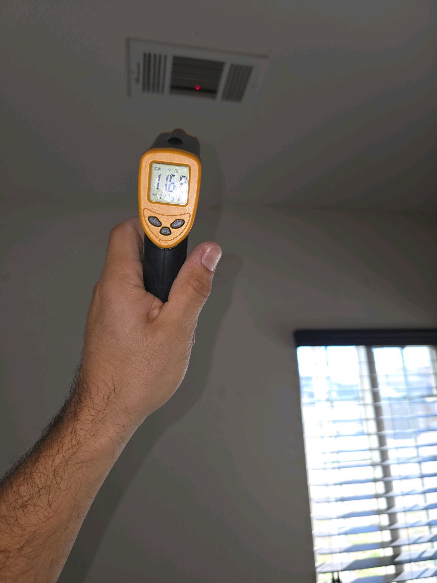 A hand holding an infrared thermometer pointing at a ceiling air vent, displaying a temperature reading of 74.6 degrees.