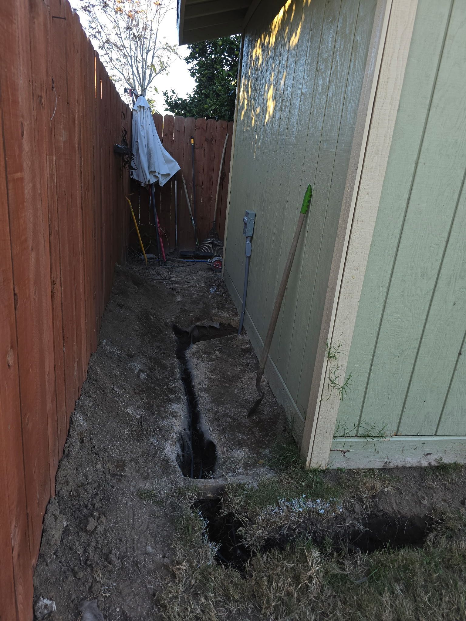 A narrow dirt trench dug along the side of a beige building next to a wooden fence.