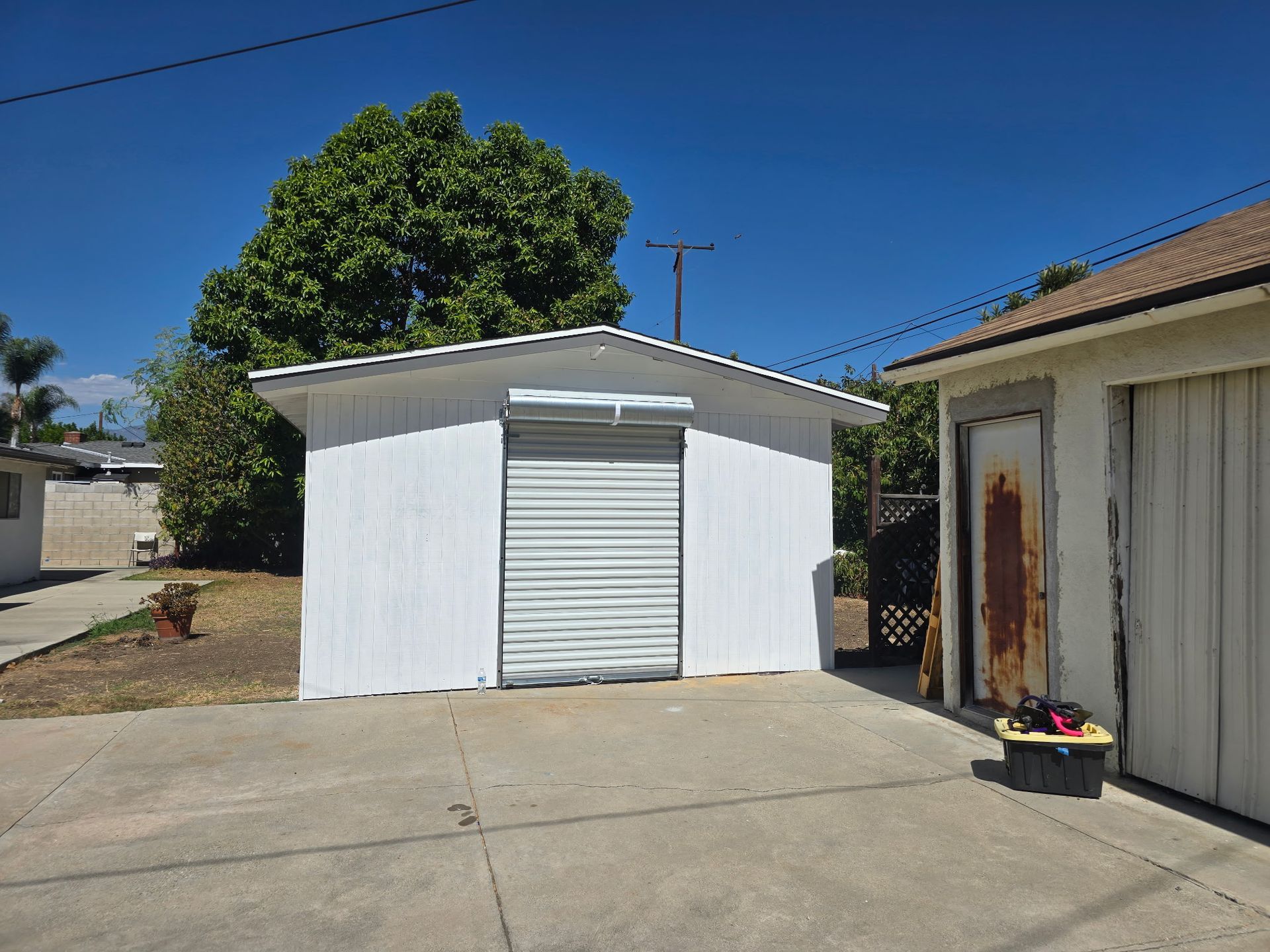 White detached garage with a roll-up door in a sunny driveway beside a house