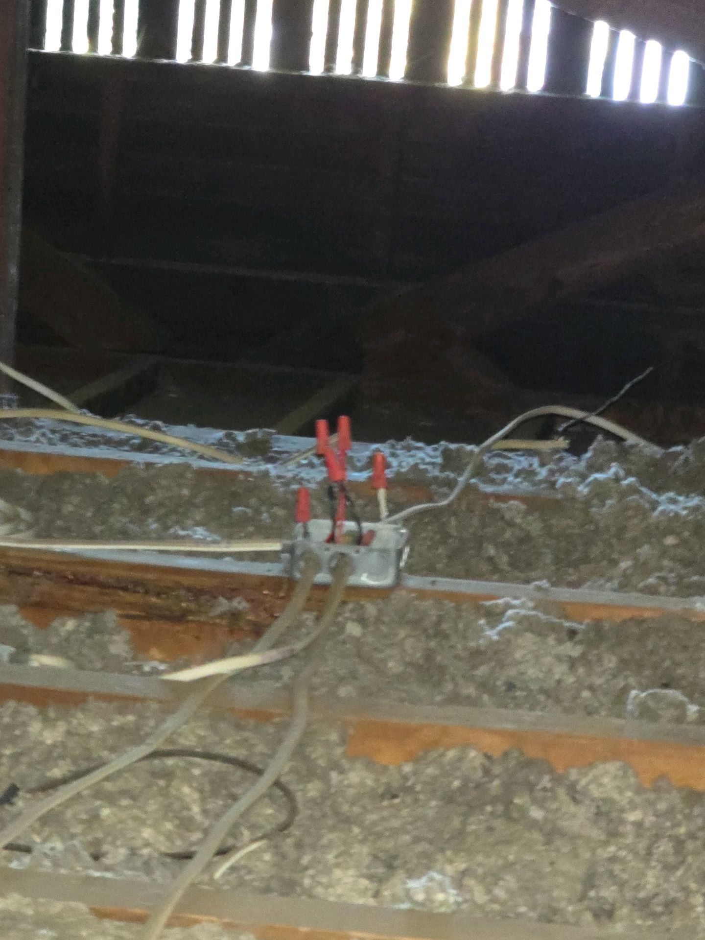A metal junction box with exposed electrical wires and red wire nuts sits in a dusty, dark attic with ceiling joists.